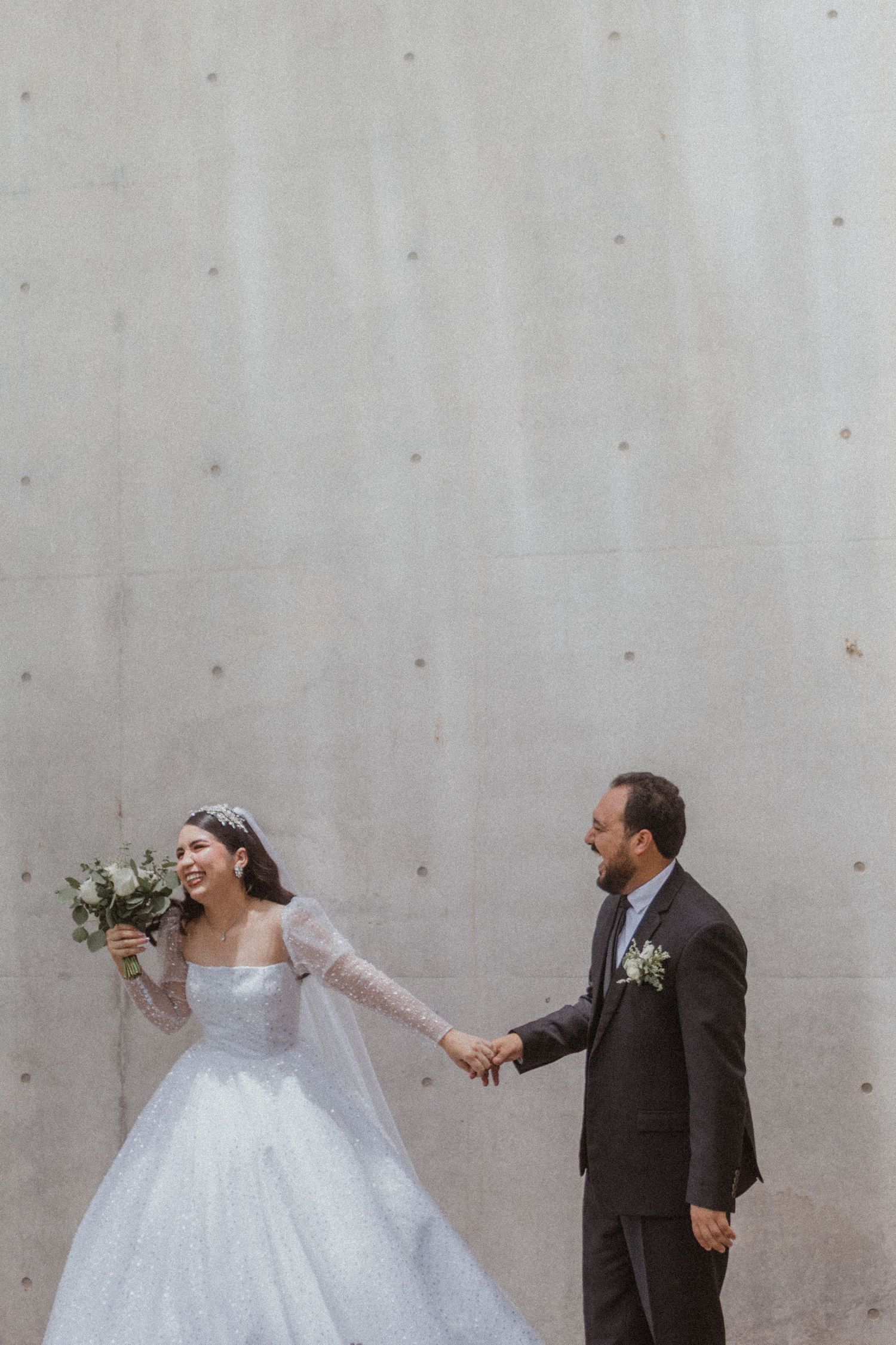 Couple in wedding attire posing playfully against a concrete wall outdoors.