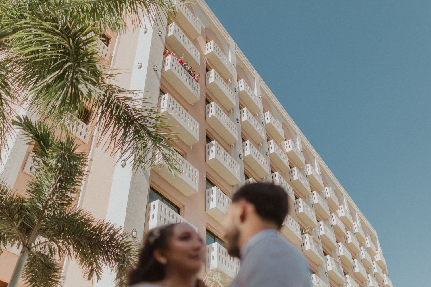 Wedding couple posing against modern hotel building with palm trees and blue sky.