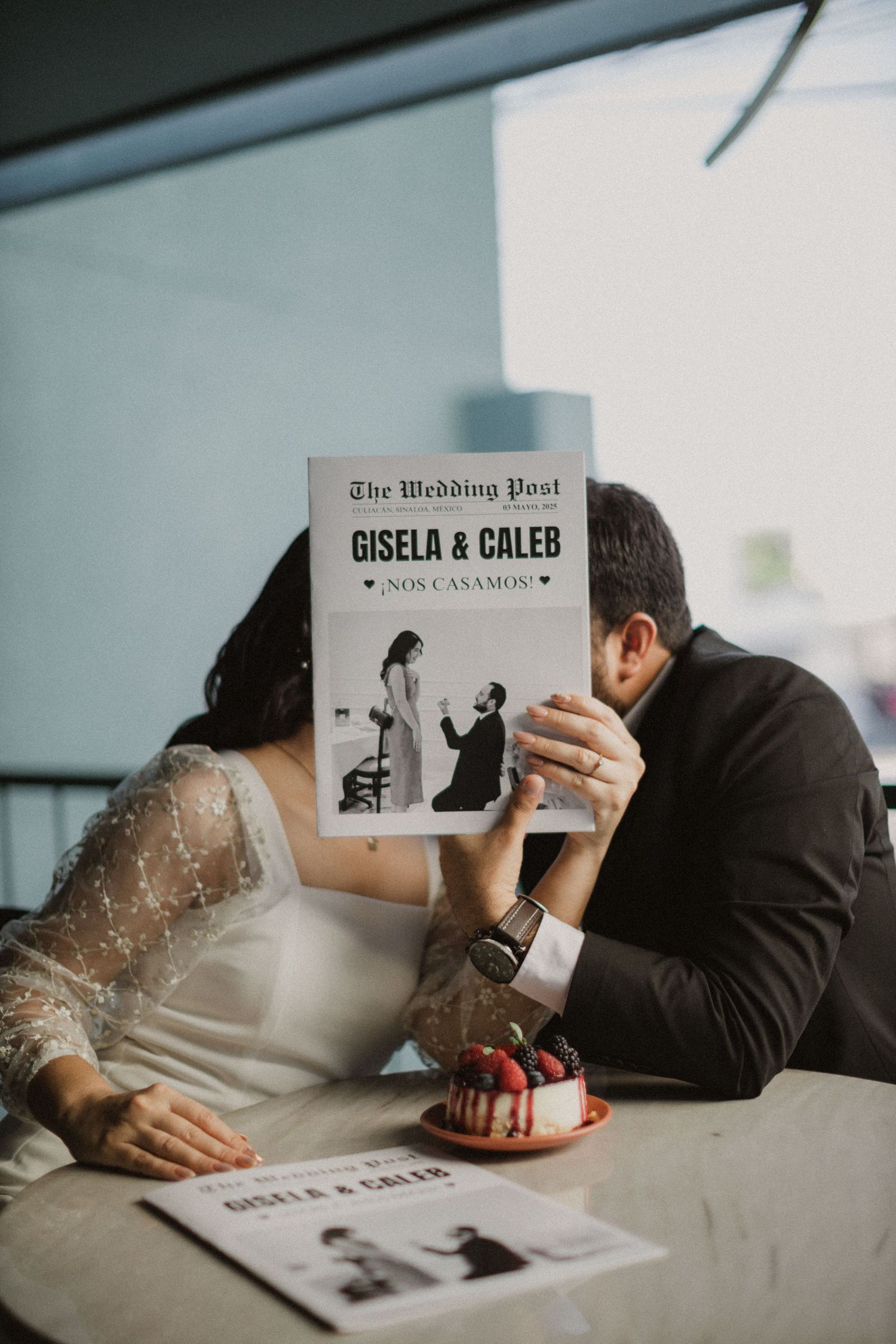 Couple shares intimate moment behind wedding newspaper at table with dessert.