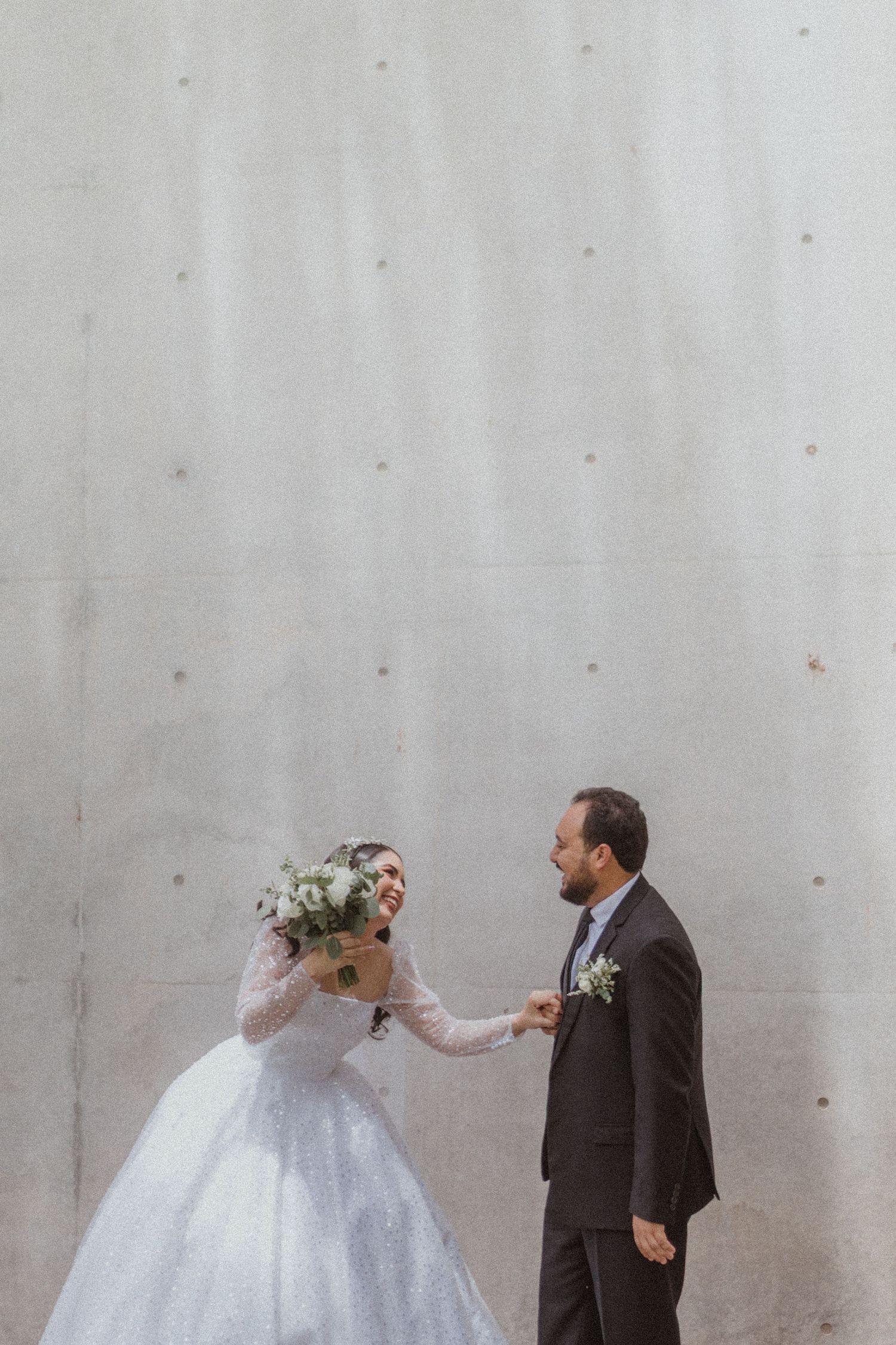 Wedding couple plays with flowers during their photo session against a minimalist concrete wall.