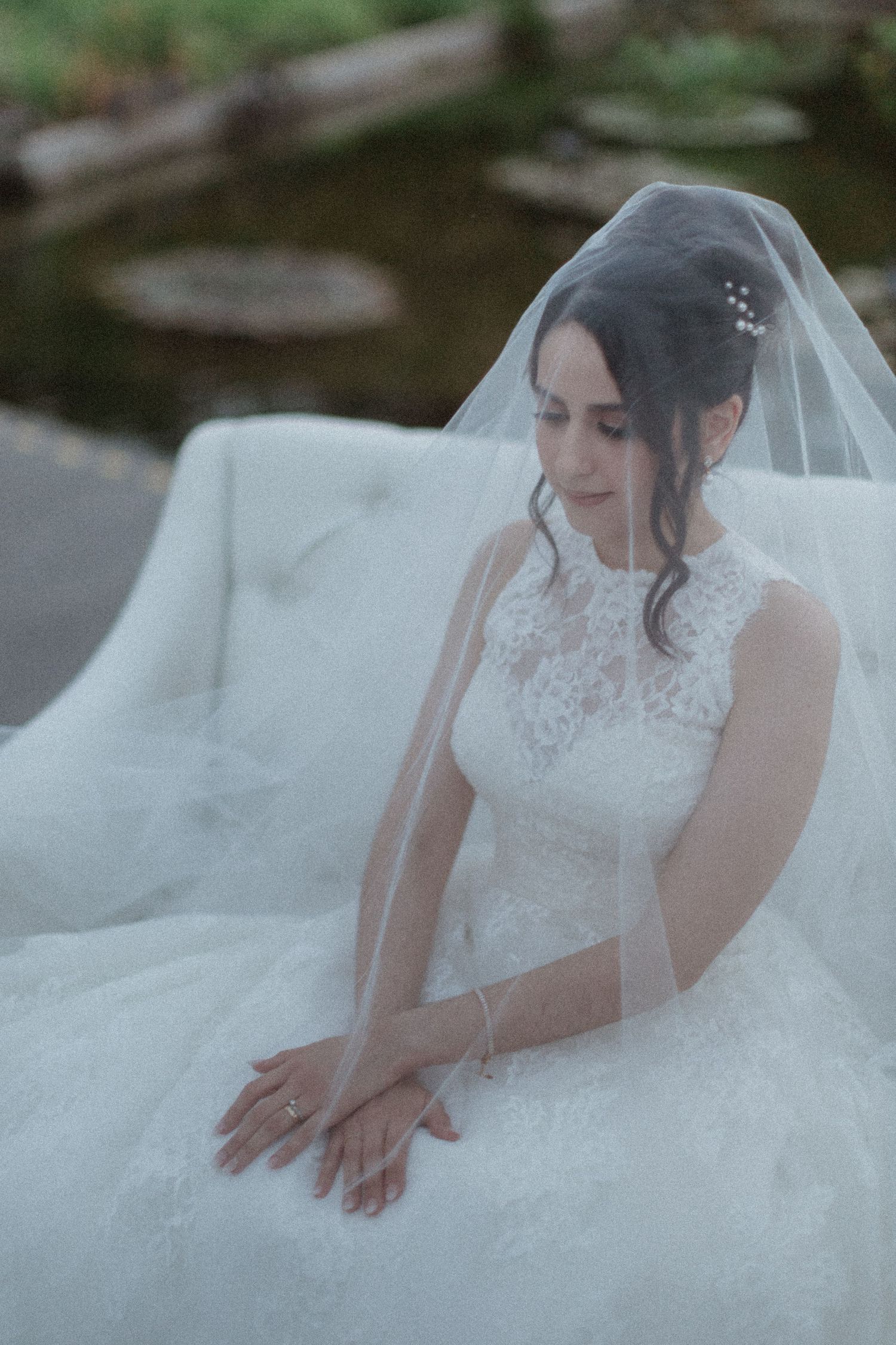 A bride in a lace wedding gown and veil poses elegantly against a nature backdrop.