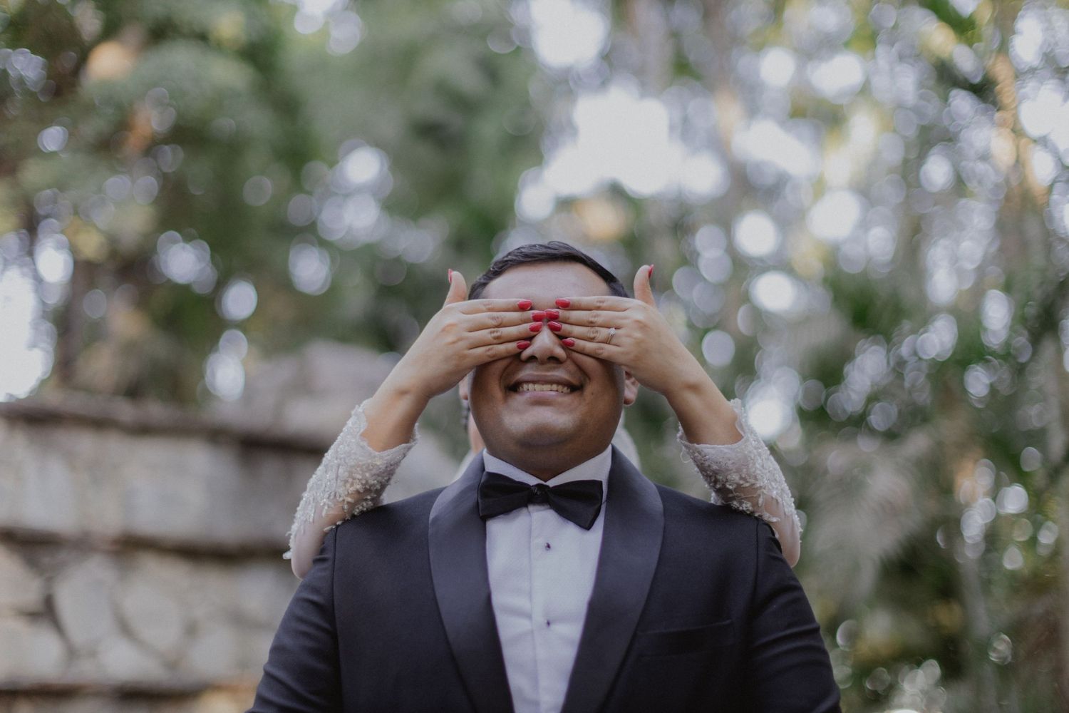 A groom in black tuxedo playfully covers his eyes during a wedding photoshoot.