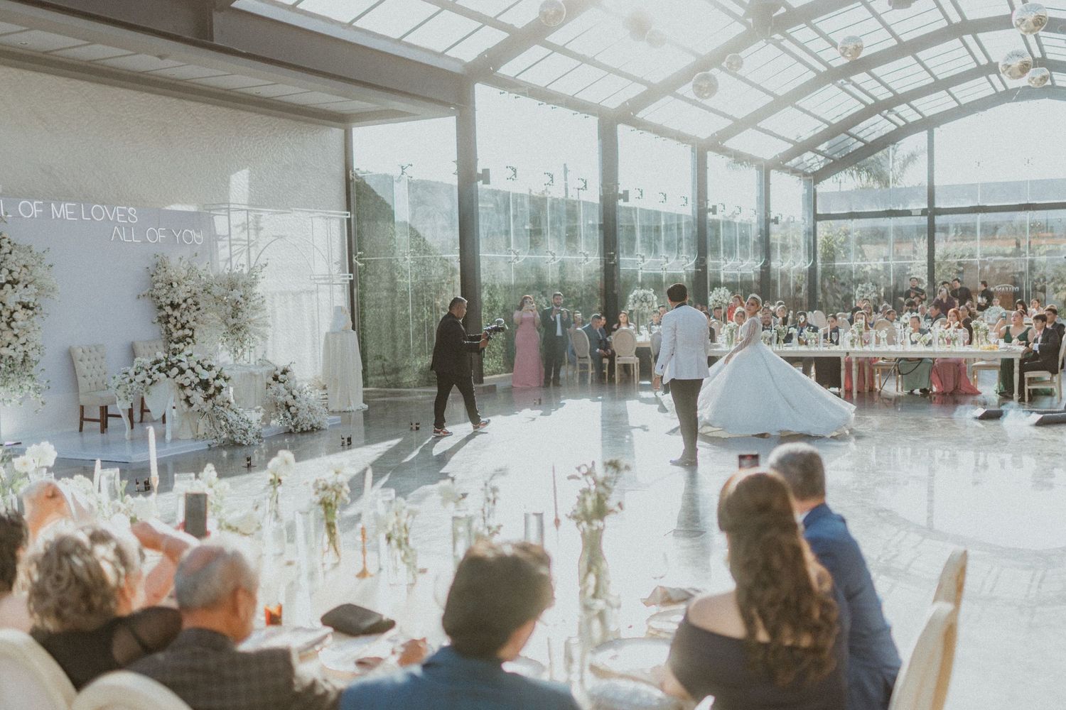 Wedding guests gather in a bright sunlit greenhouse venue with glass ceiling and elegant table settings for a reception celebration.