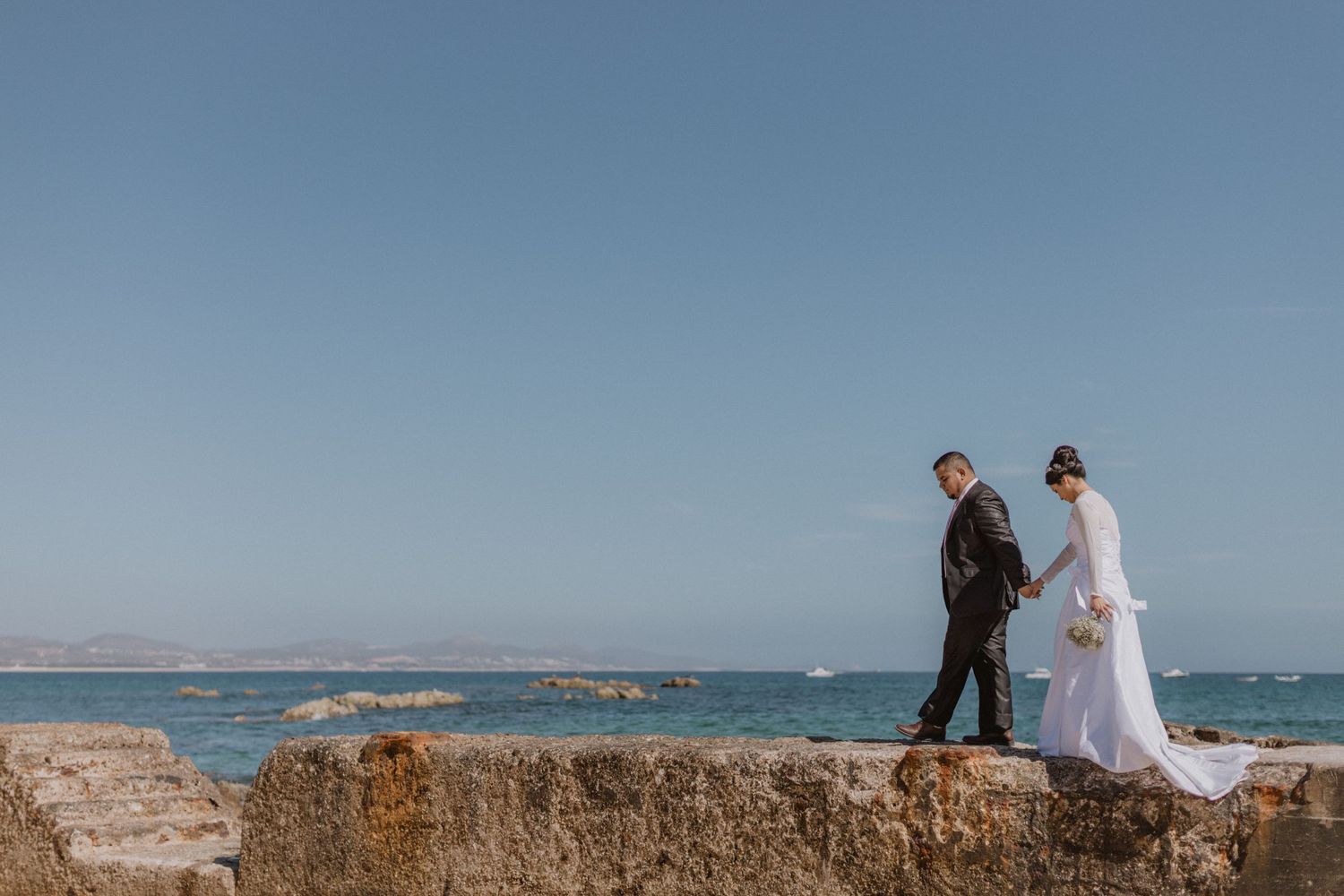 Couple in wedding attire walking on rocky coastal landscape.