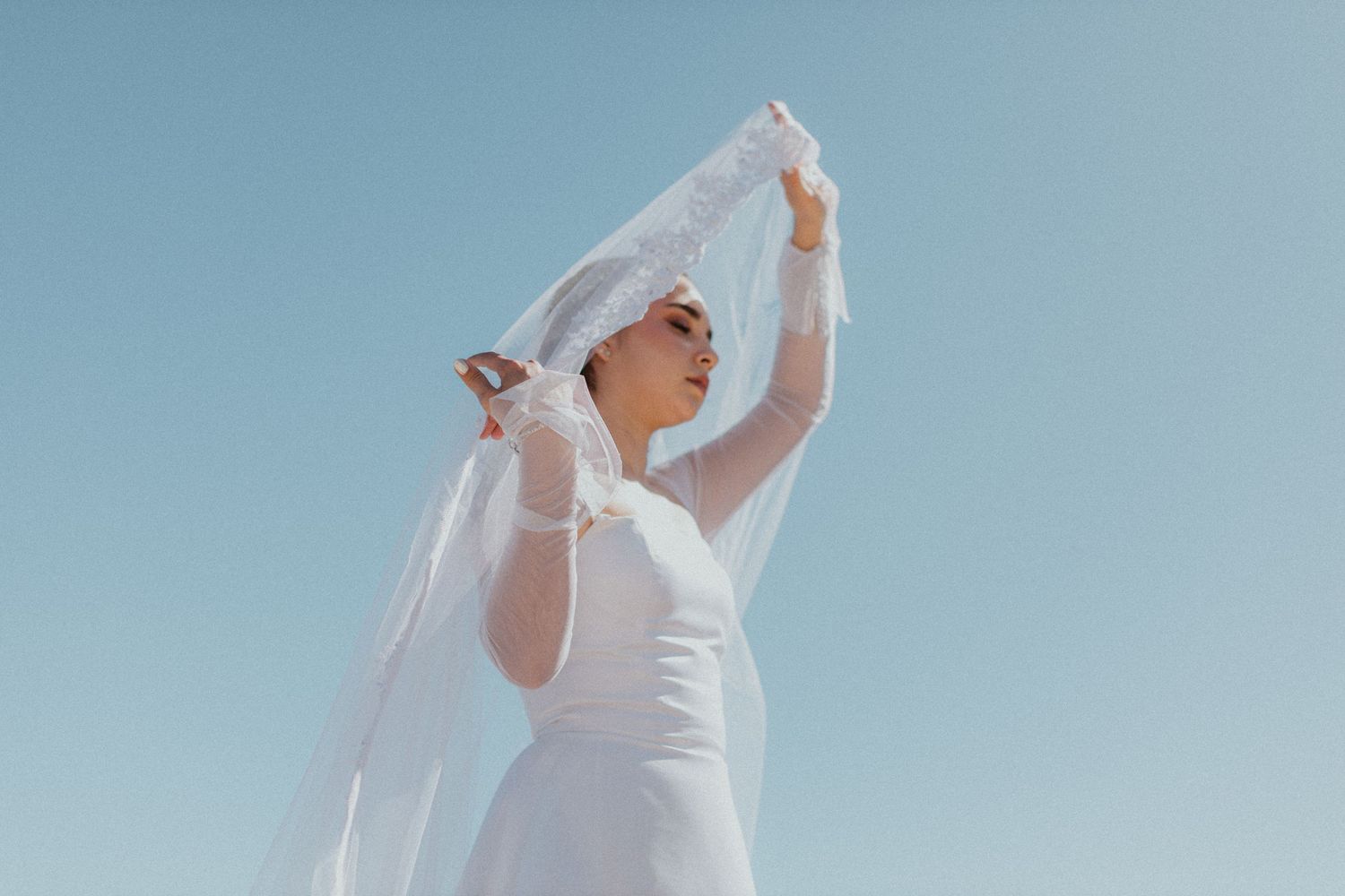 A bride in white dress holds her veil against bright blue sky.