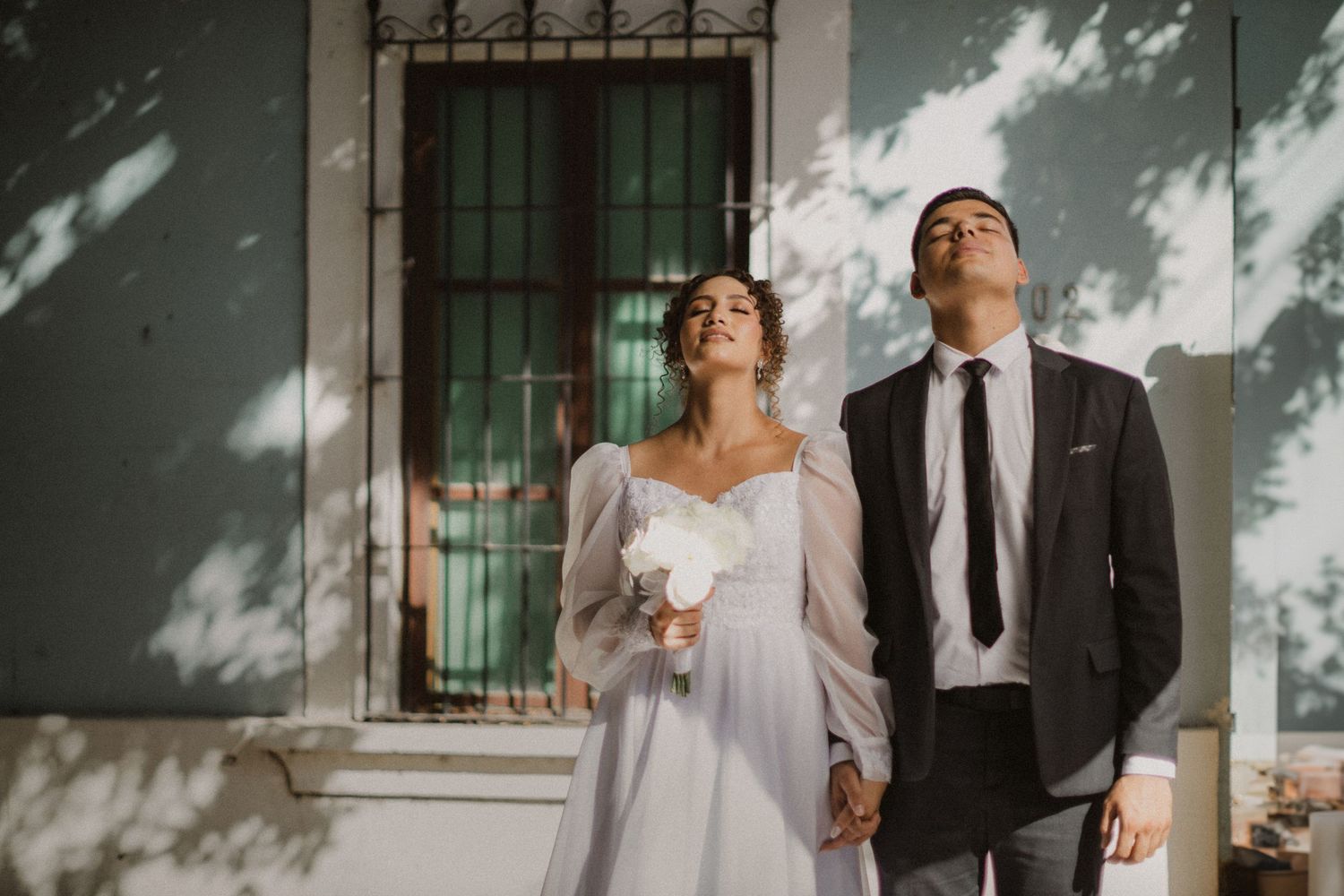 A couple in wedding attire standing together against a white wall with dappled sunlight and shadows.