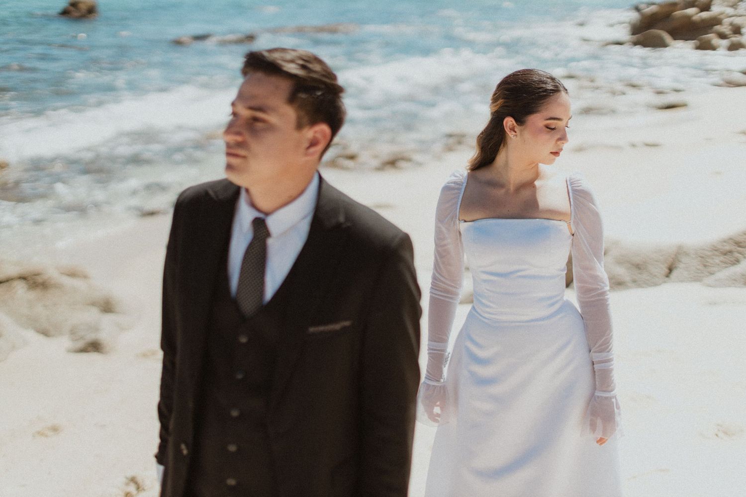 Elegant couple in wedding attire standing on beach with ocean view.