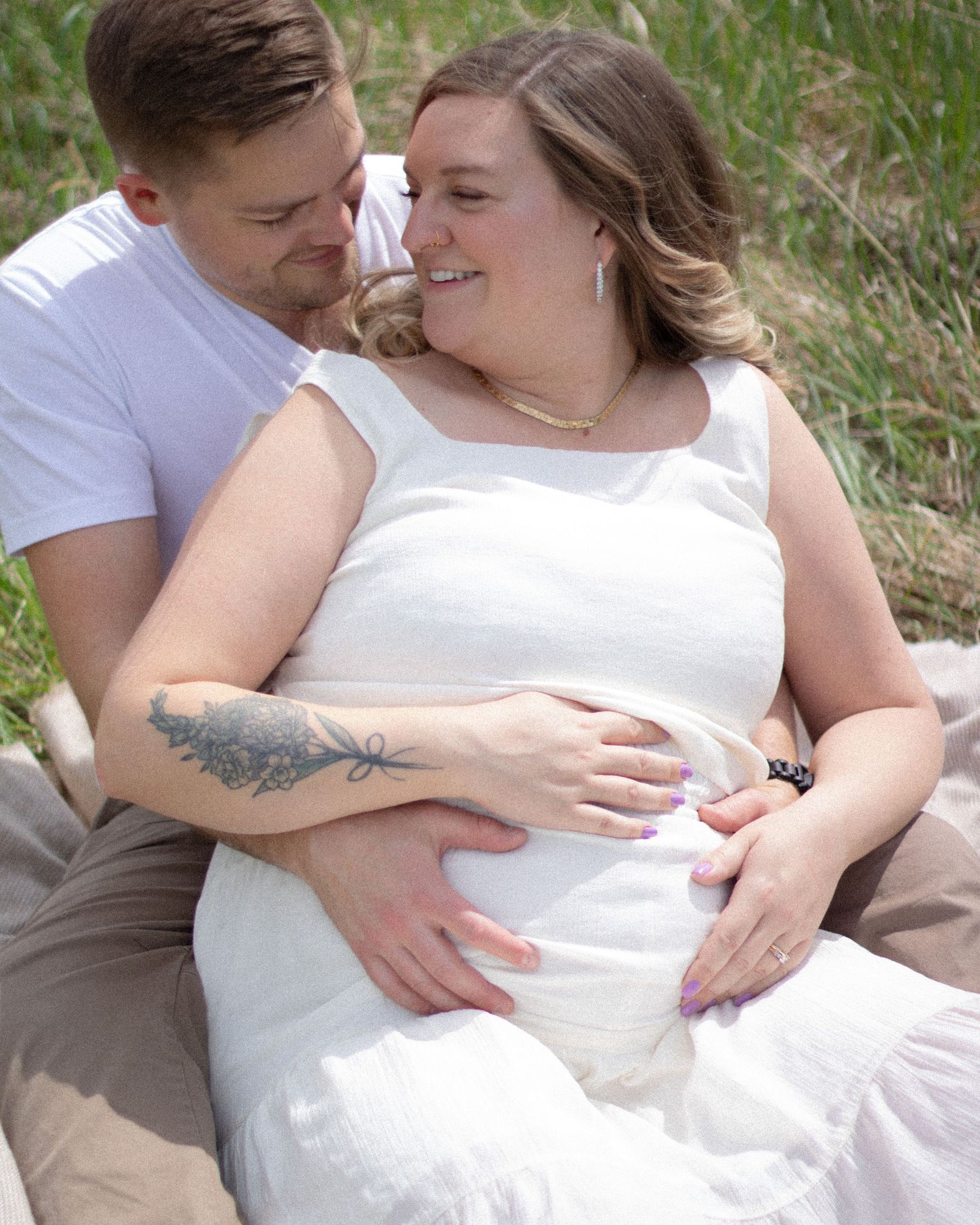 Couple sitting outdoors in a maternity photo shoot, man embracing pregnant woman in white dress.