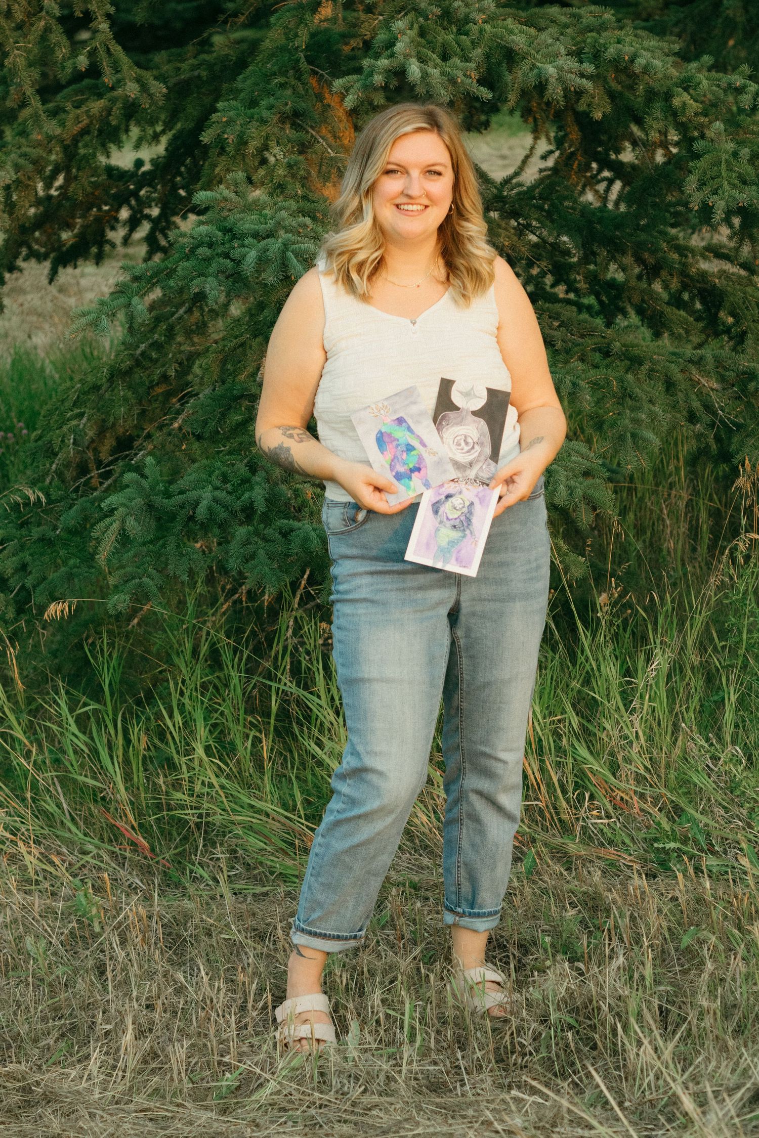Smiling woman in white top and jeans holding colorful artwork while standing near evergreen trees.