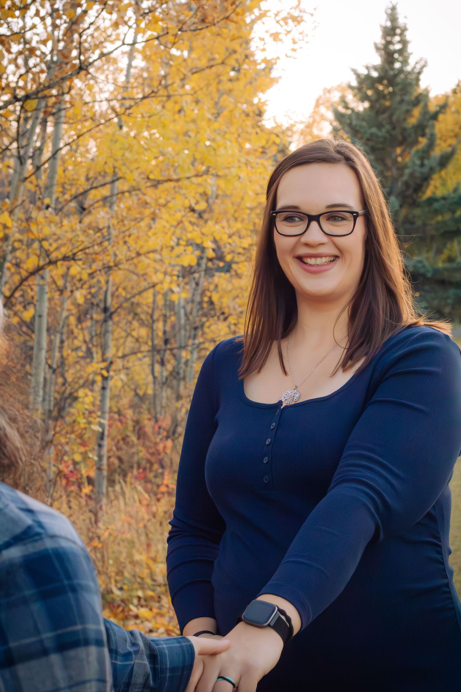 Smiling pregnant woman with glasses in navy blue dress among autumn yellow trees.
