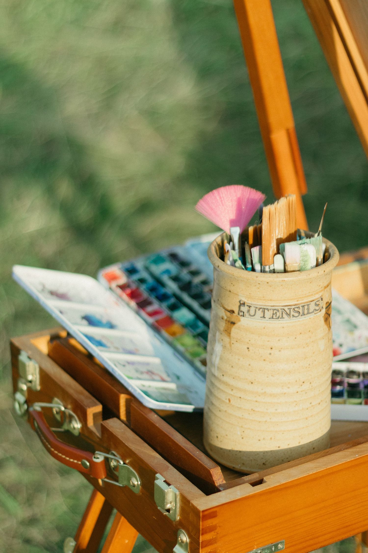 Artist outdoor easel with ceramic utensil holder, brushes, and watercolor palette on wooden drawer.