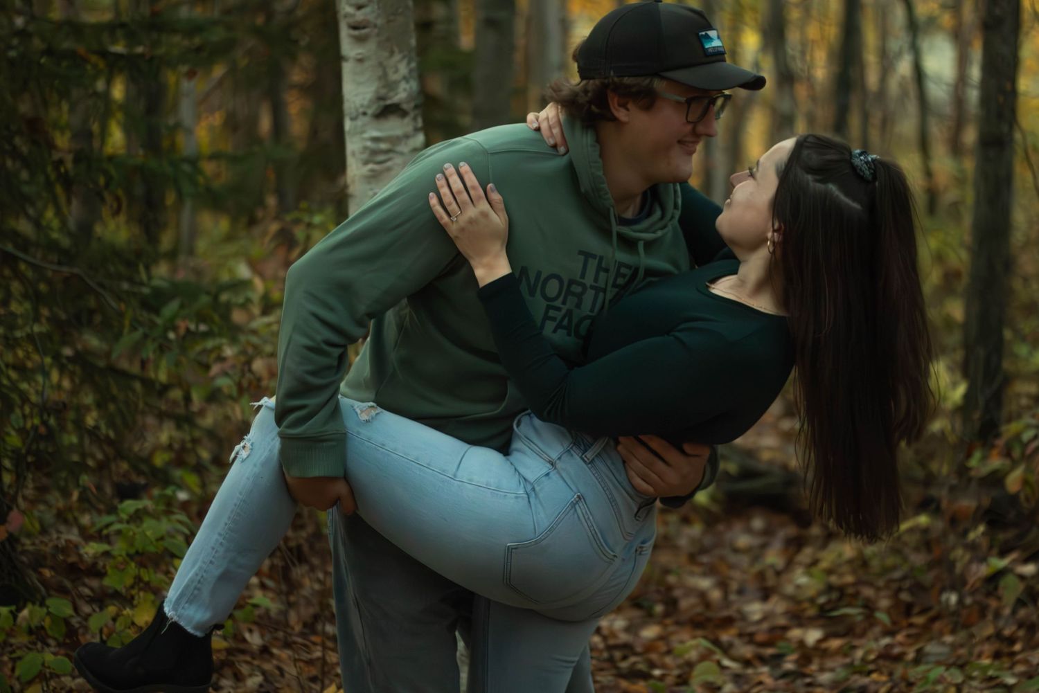 A couple embraces and kisses as the man lifts the woman in light blue jeans in an autumn forest.