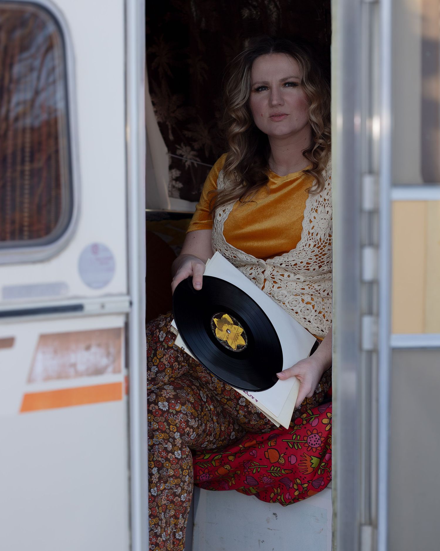 Vintage-styled woman sitting in a retro caravan doorway holding a vinyl record in boho attire.