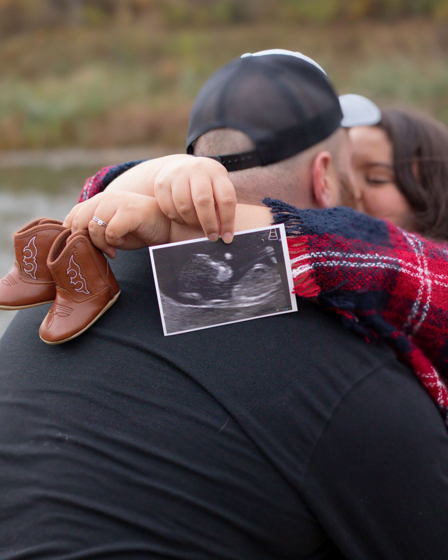 Couple embracing outdoors holding ultrasound photo and tiny cowboy boots, announcing pregnancy in autumn setting.