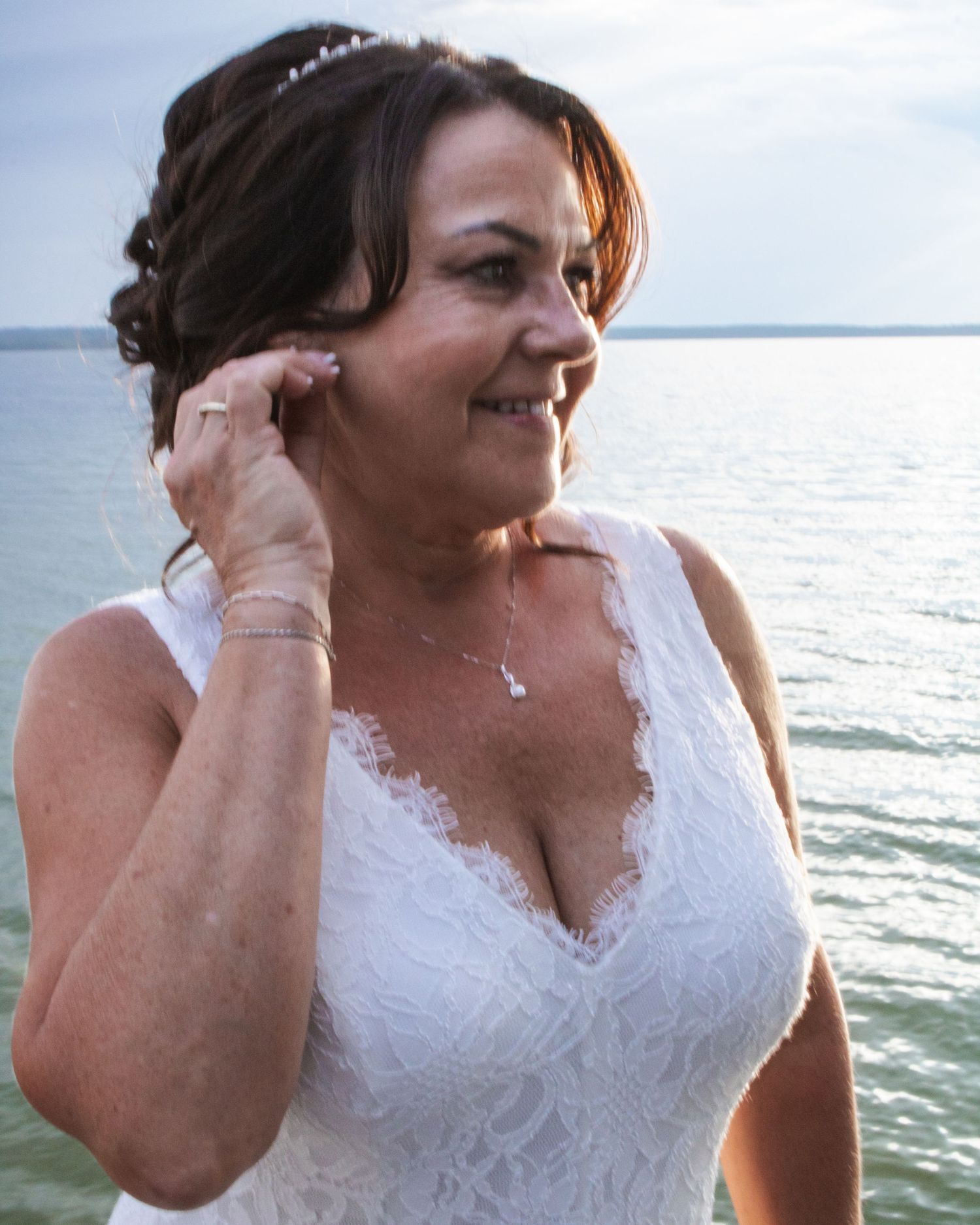 Bride in white lace V-neck dress adjusts earring while standing near sparkling blue water outdoors.