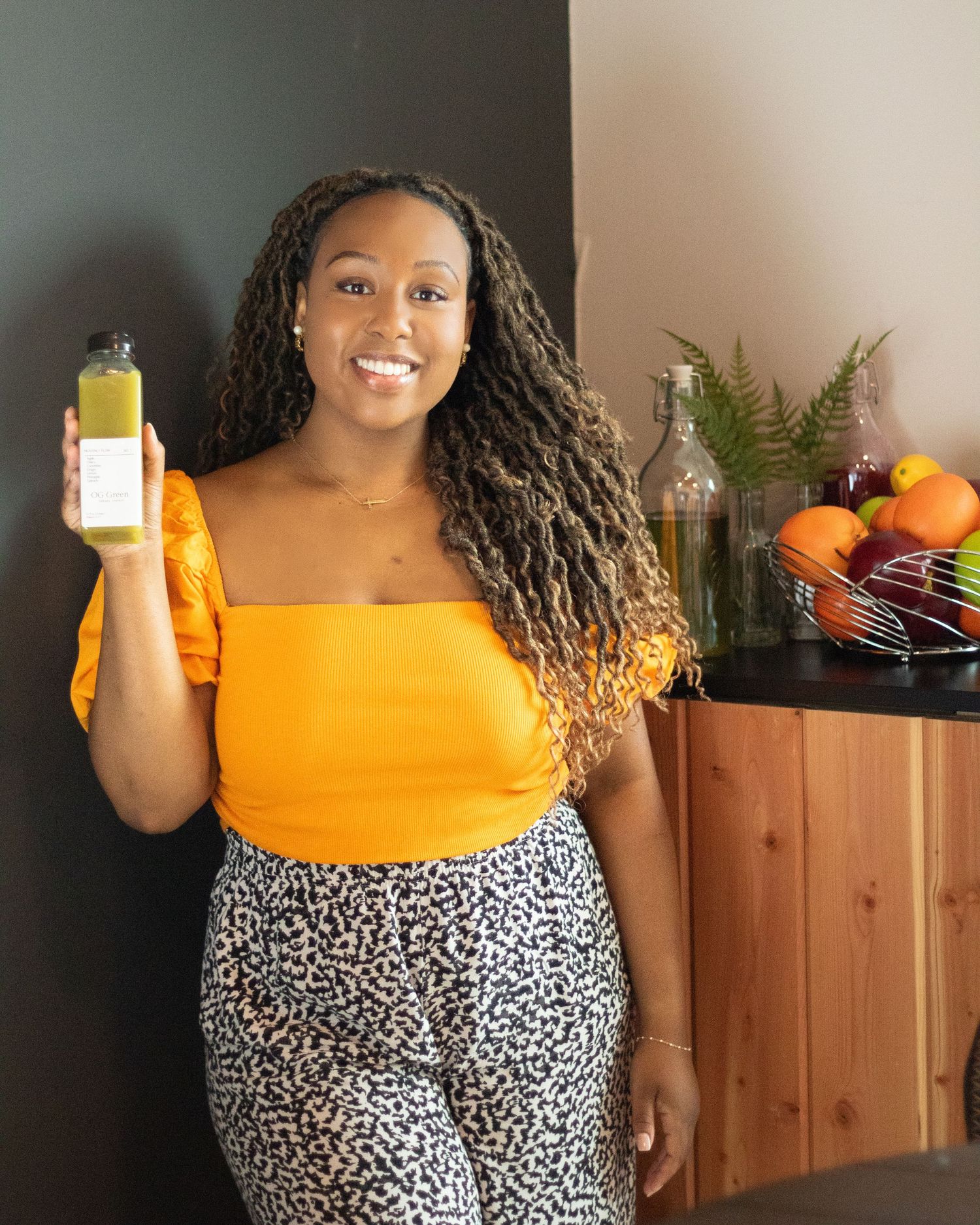Smiling woman in orange top holding a green juice bottle in a bright kitchen with fresh fruits.