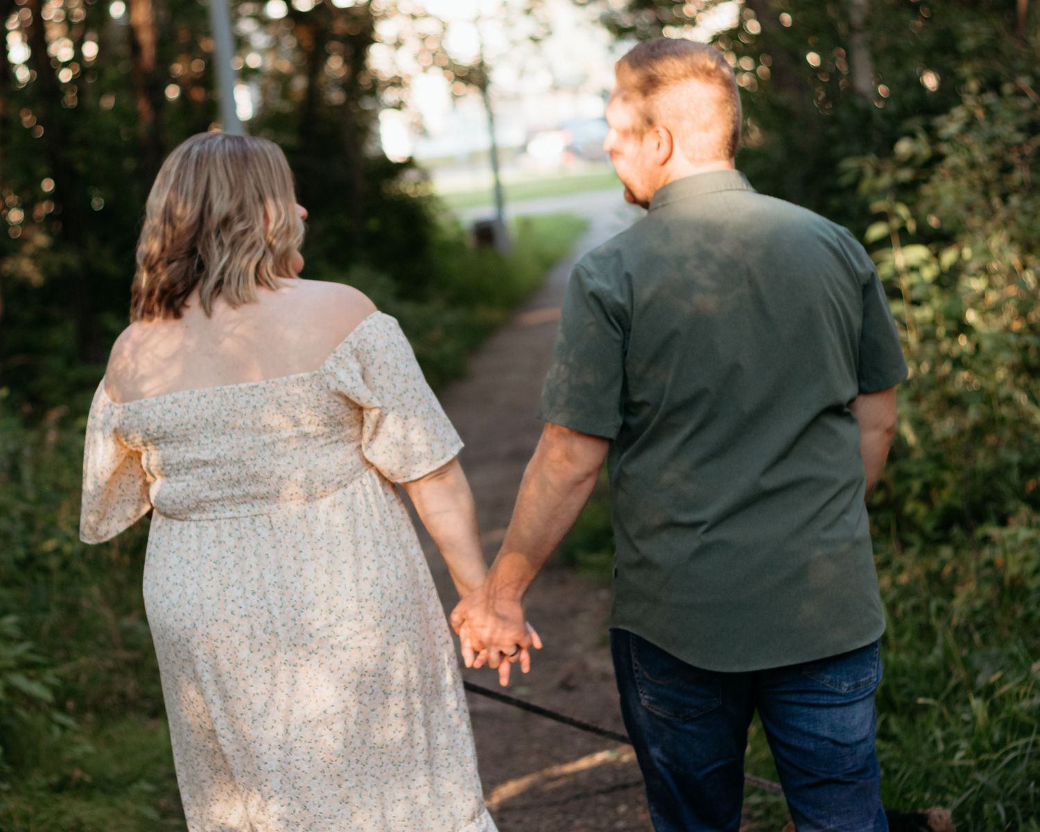 Pregnant couple walking hand in hand down sunlit garden path surrounded by trees.