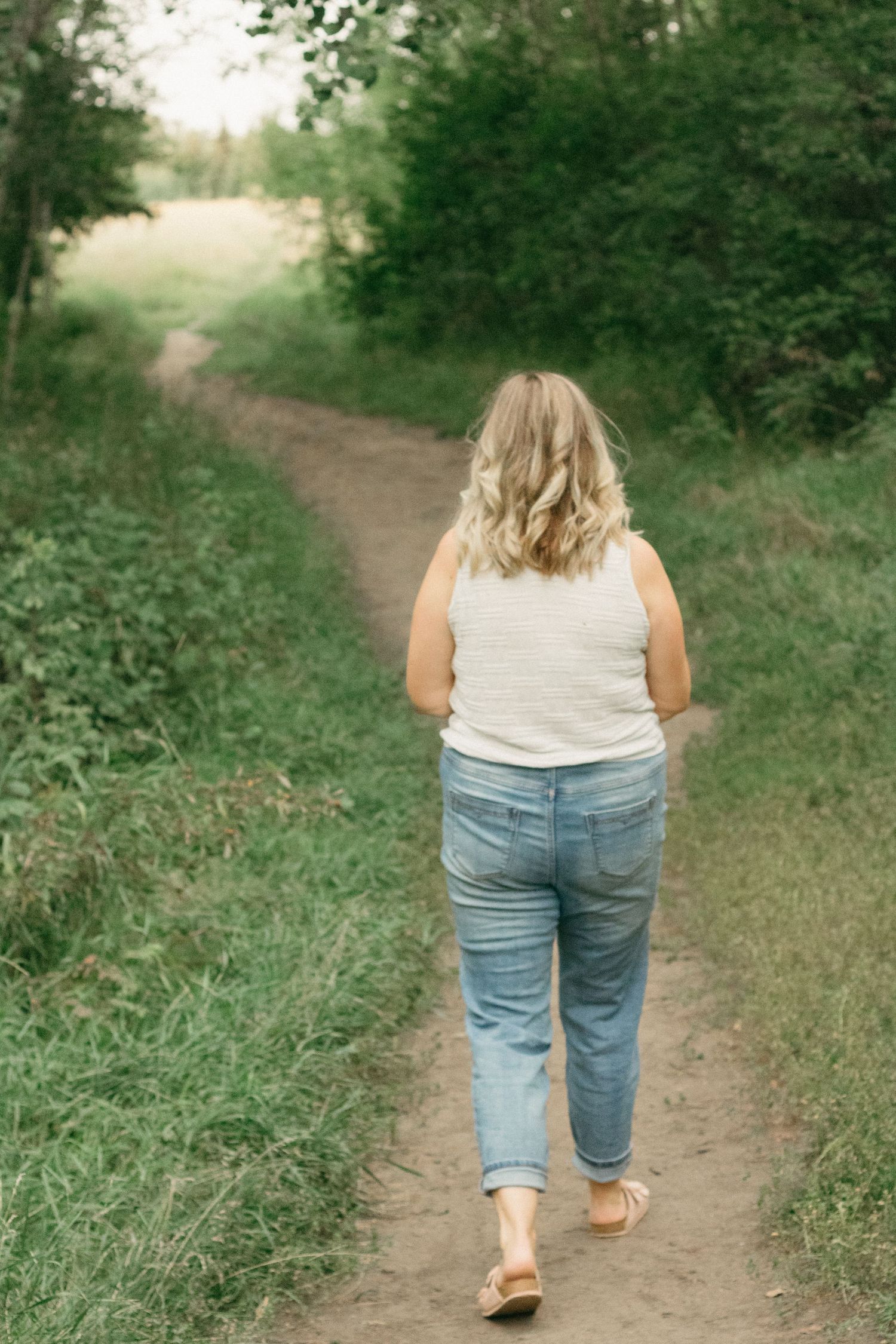 Blonde woman in white tank top and jeans walking away on a dirt path surrounded by lush greenery.