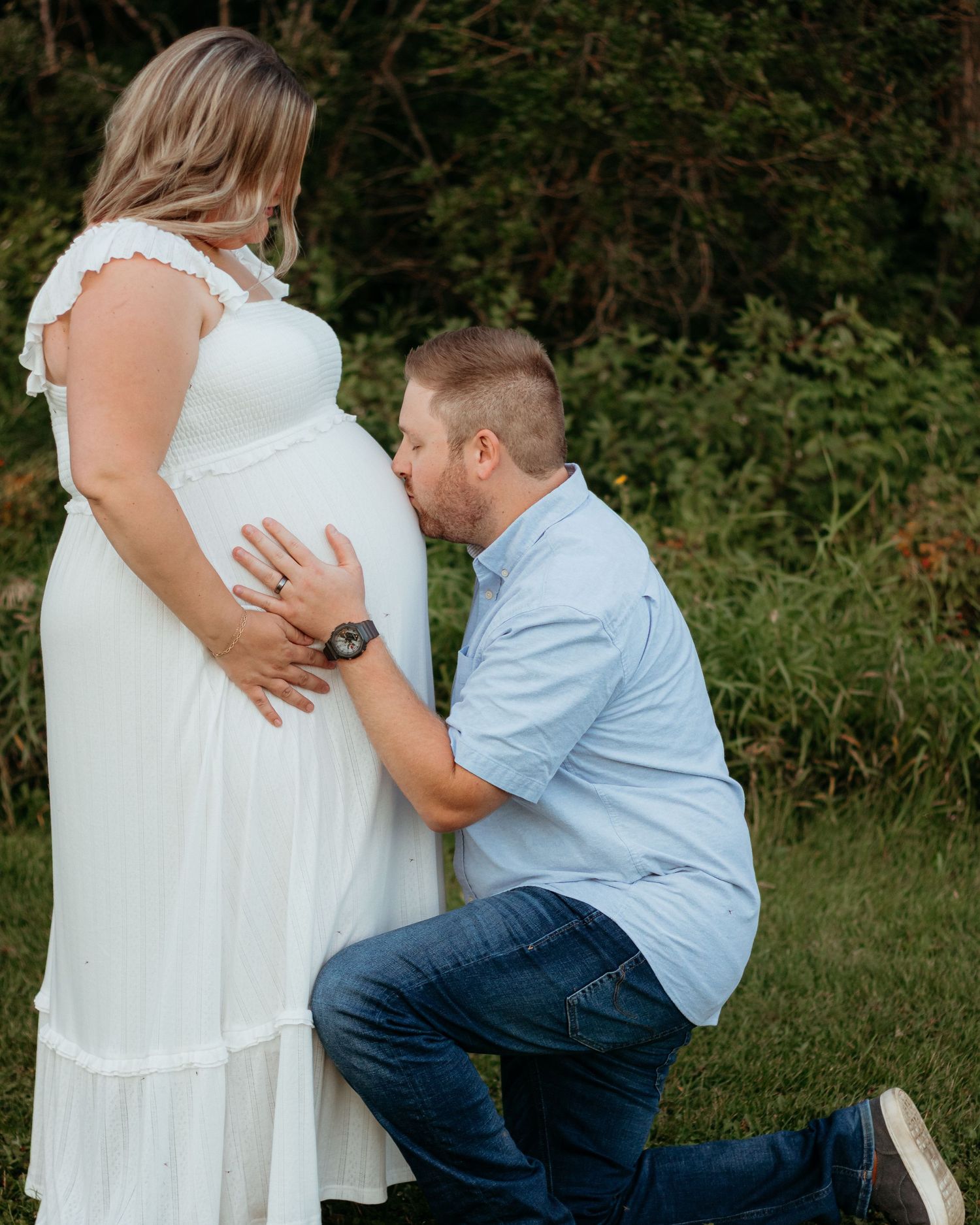 Man kneeling and kissing pregnant woman's belly outdoors, maternity photo shoot in white dress.