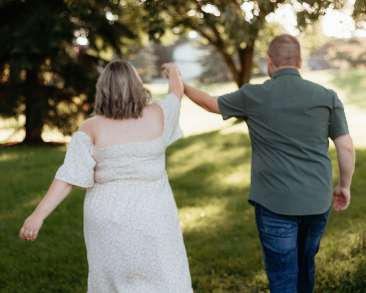 Pregnant couple walking hand in hand through a sunny green park, viewed from behind.