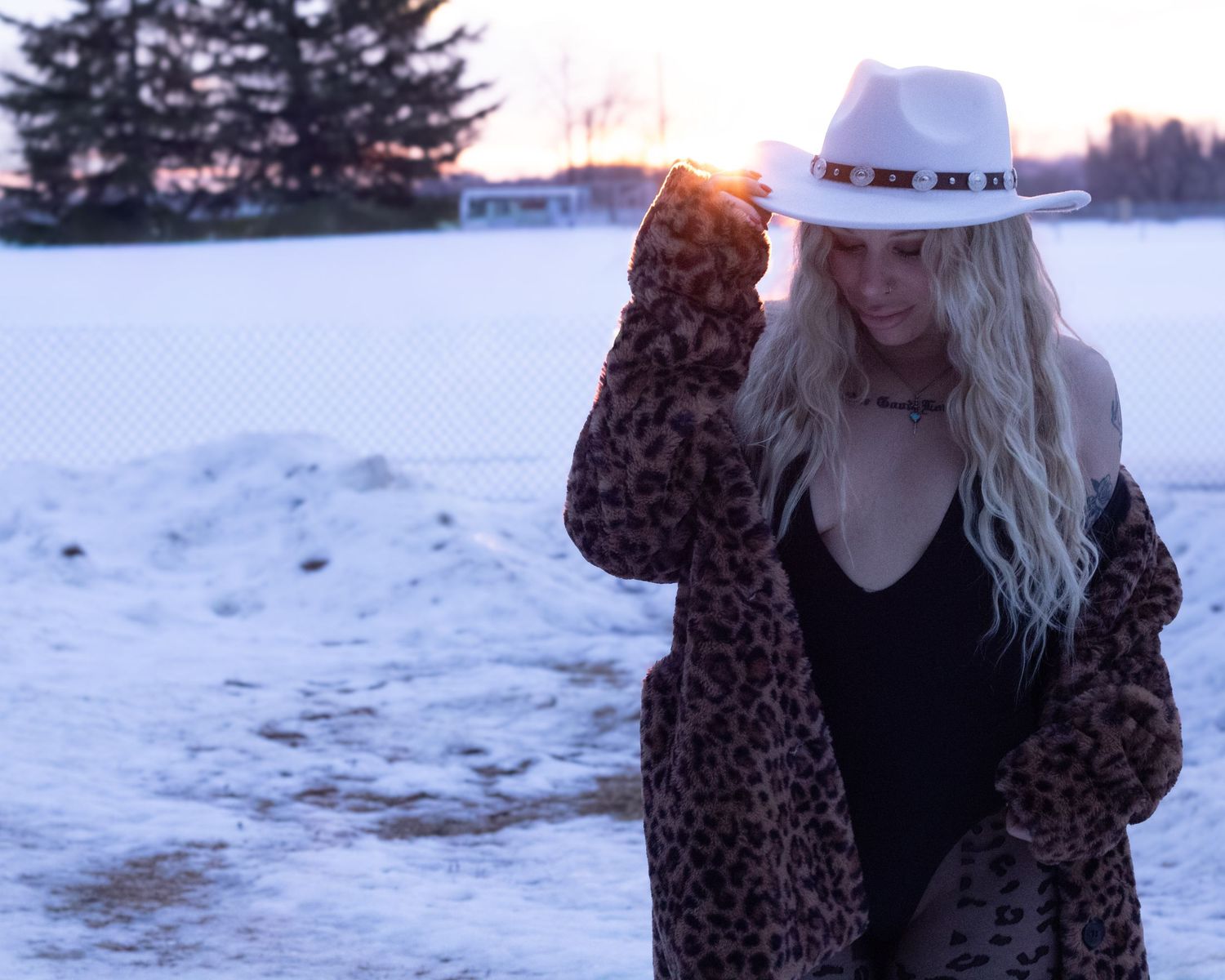 Blonde woman in leopard fur coat and white hat standing in snowy field at sunset.