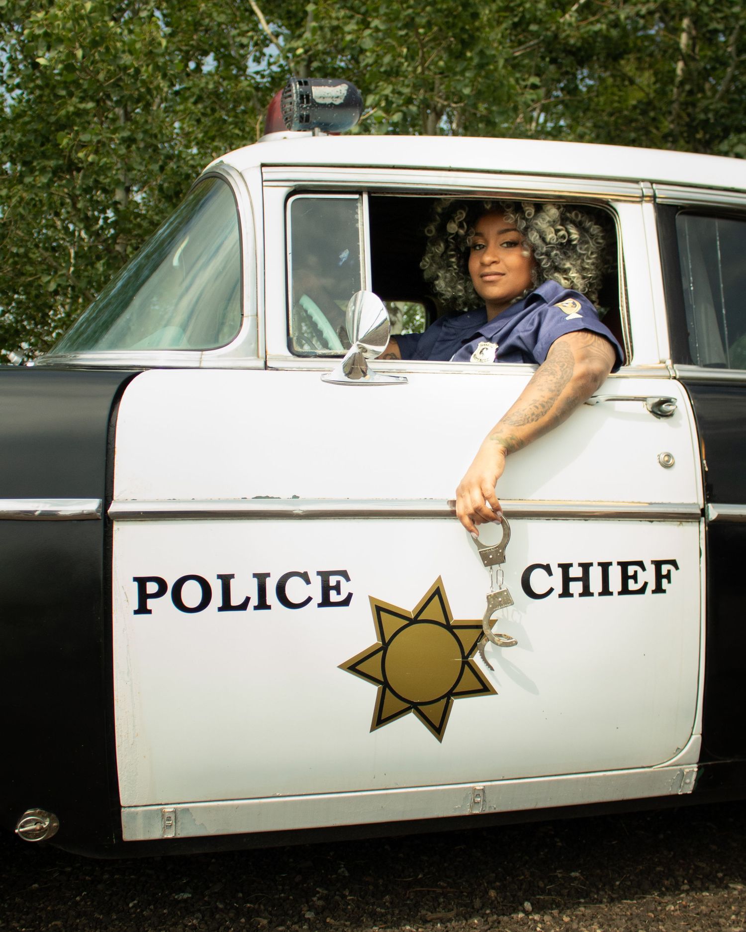 A person in a police uniform leans from a vintage Police Chief car window, dangling handcuffs.