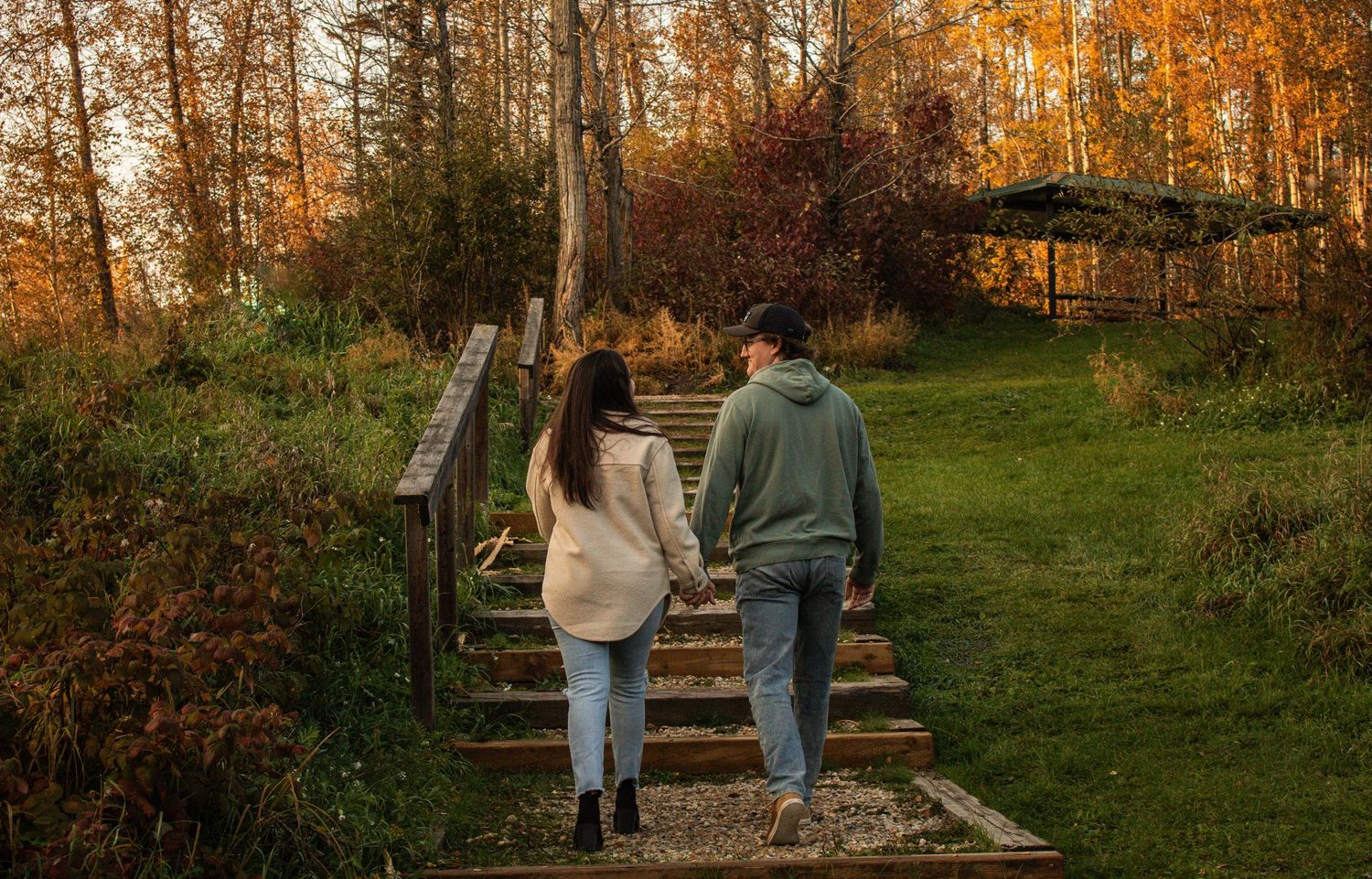 Couple holding hands walking up wooden steps through a vibrant autumn forest landscape.
