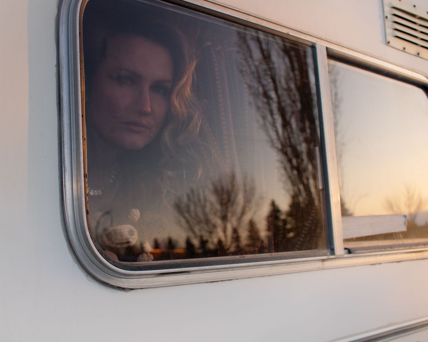 Person gazing through a vintage camper window with trees reflected in the golden sunset light.