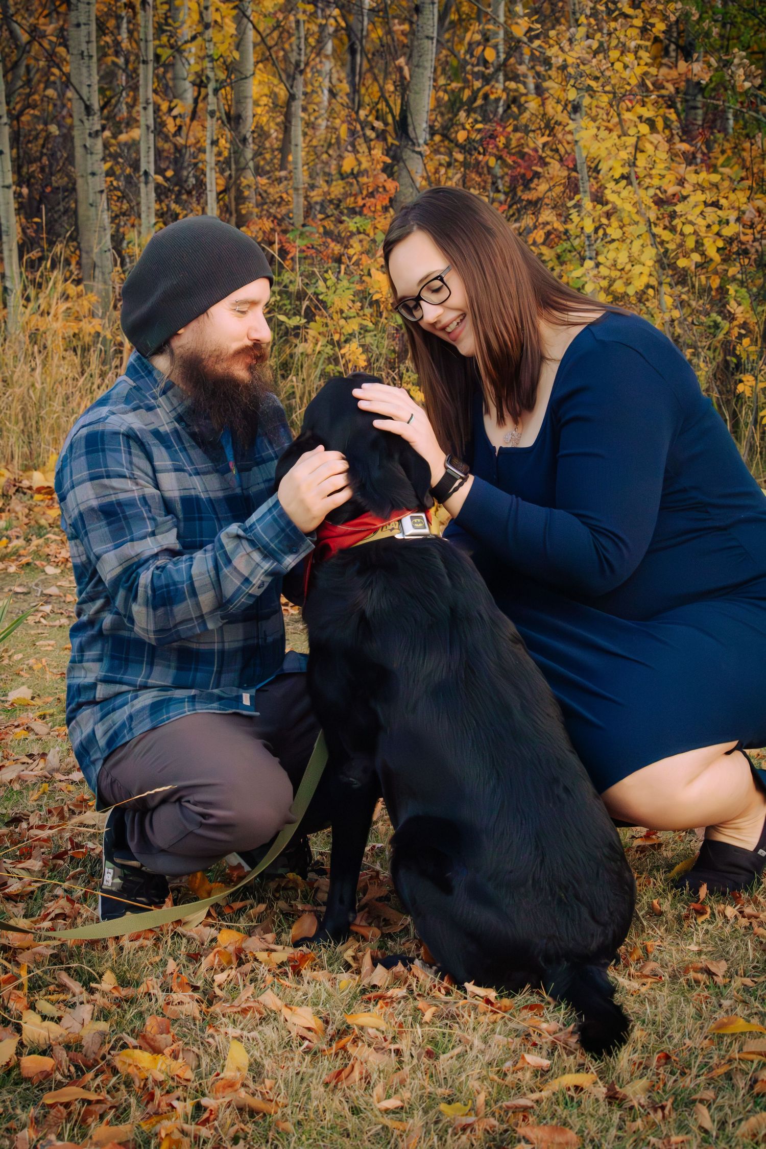 Couple crouching with black Labrador dog among fallen autumn leaves in wooded area.