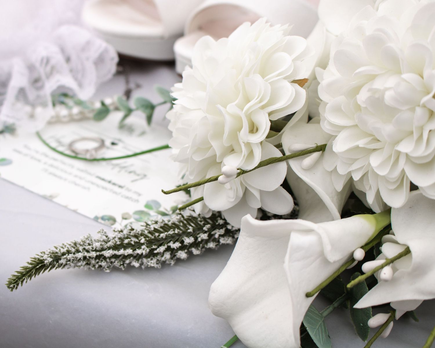 White wedding bouquet with dahlias and greenery beside wedding invitation and bridal shoes.
