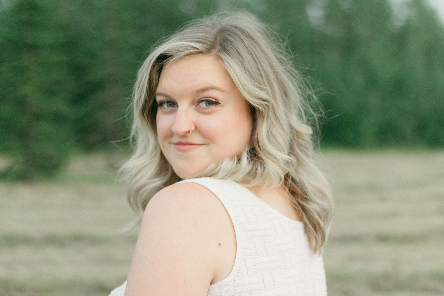 Smiling blonde woman in white top looking back over her shoulder with pine trees behind her.