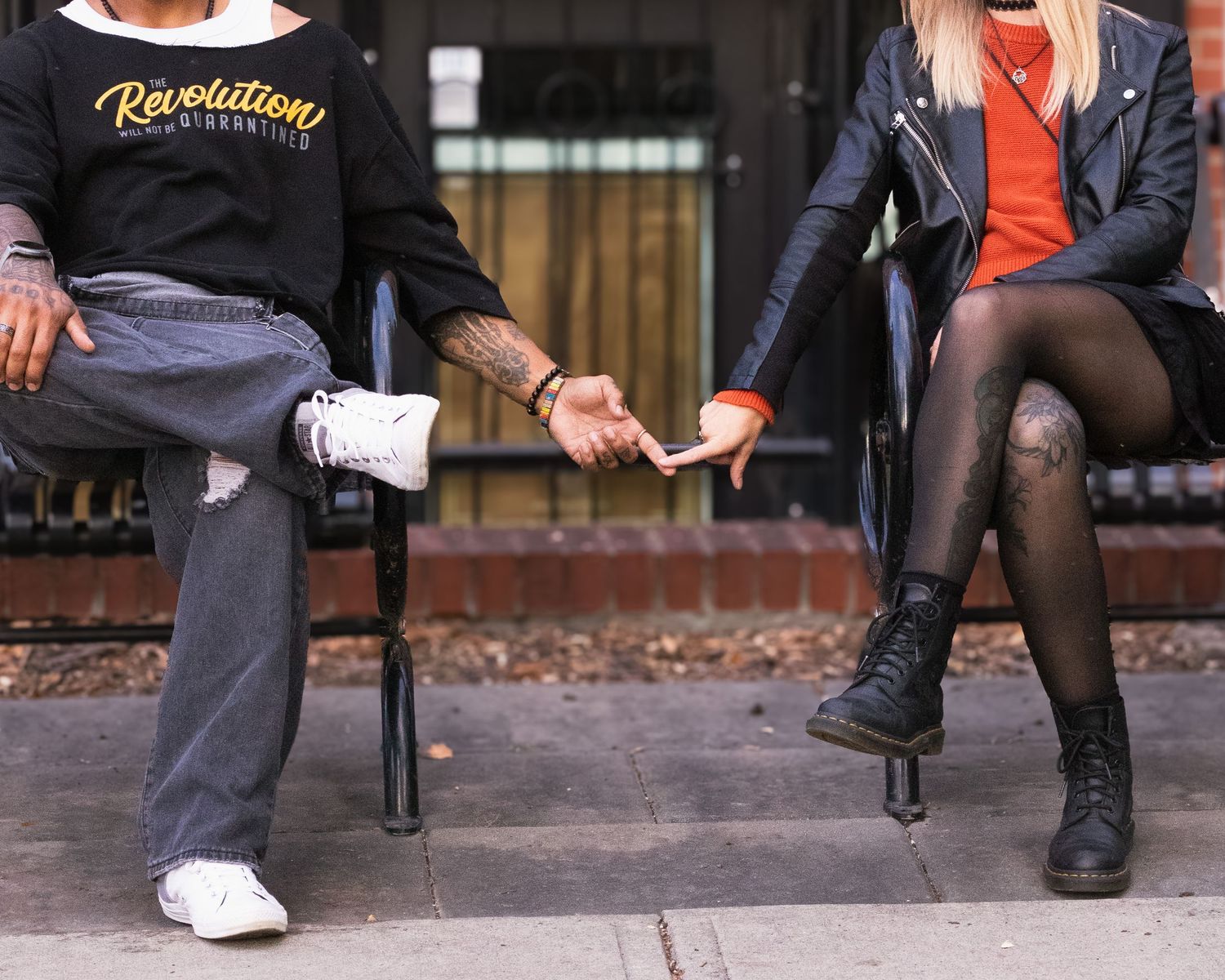 Couple sitting apart on chairs holding hands, man in gray jeans and sneakers, woman in black leather jacket and boots.