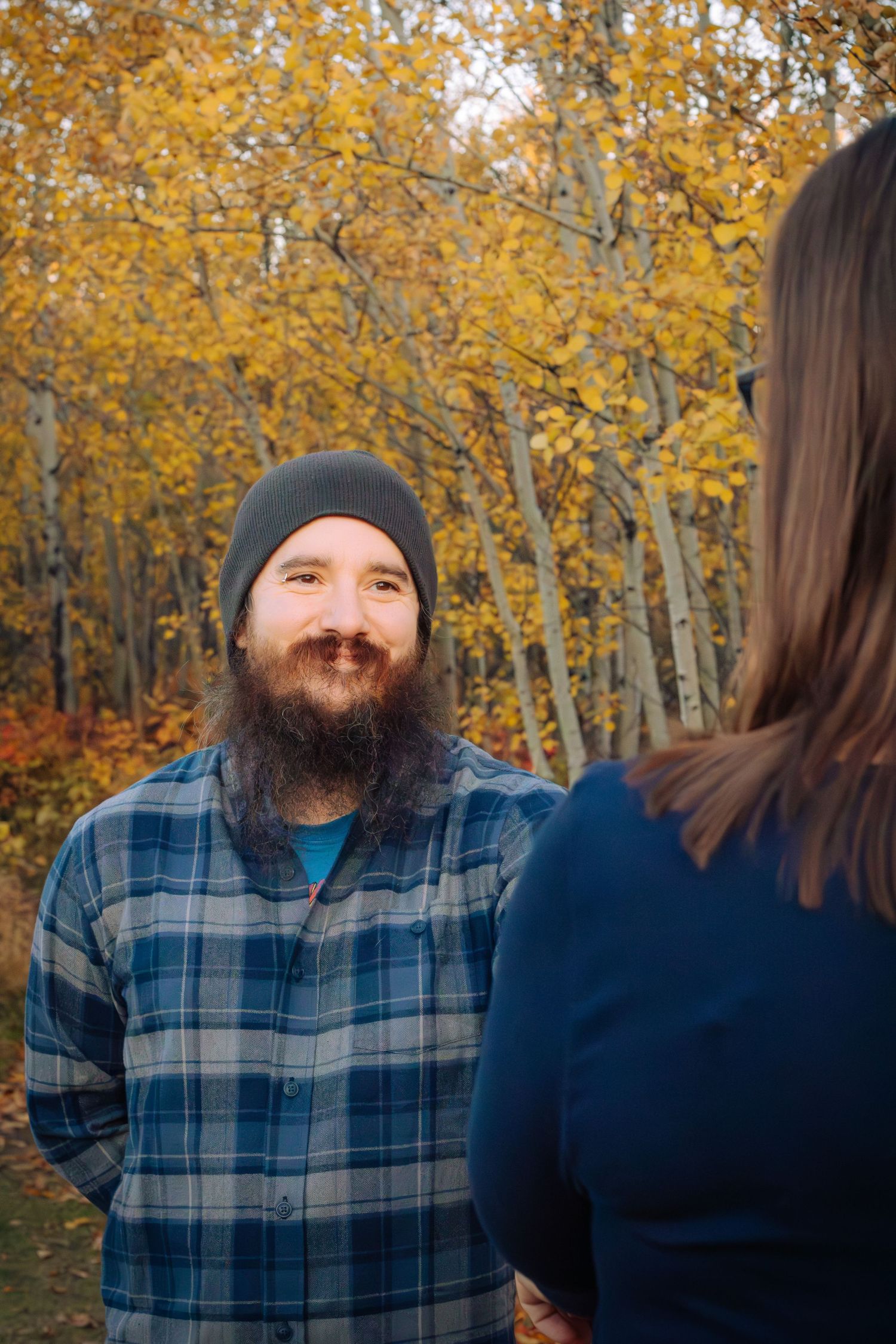 Bearded man in plaid shirt and beanie smiling at woman in autumn forest setting.