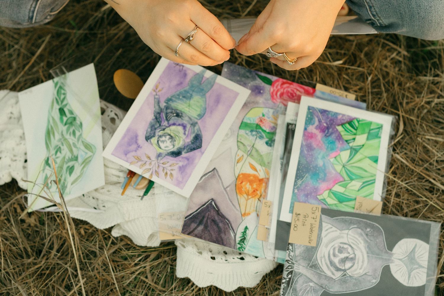 Artist reviewing colorful watercolor paintings laid out on dry grass outdoors.