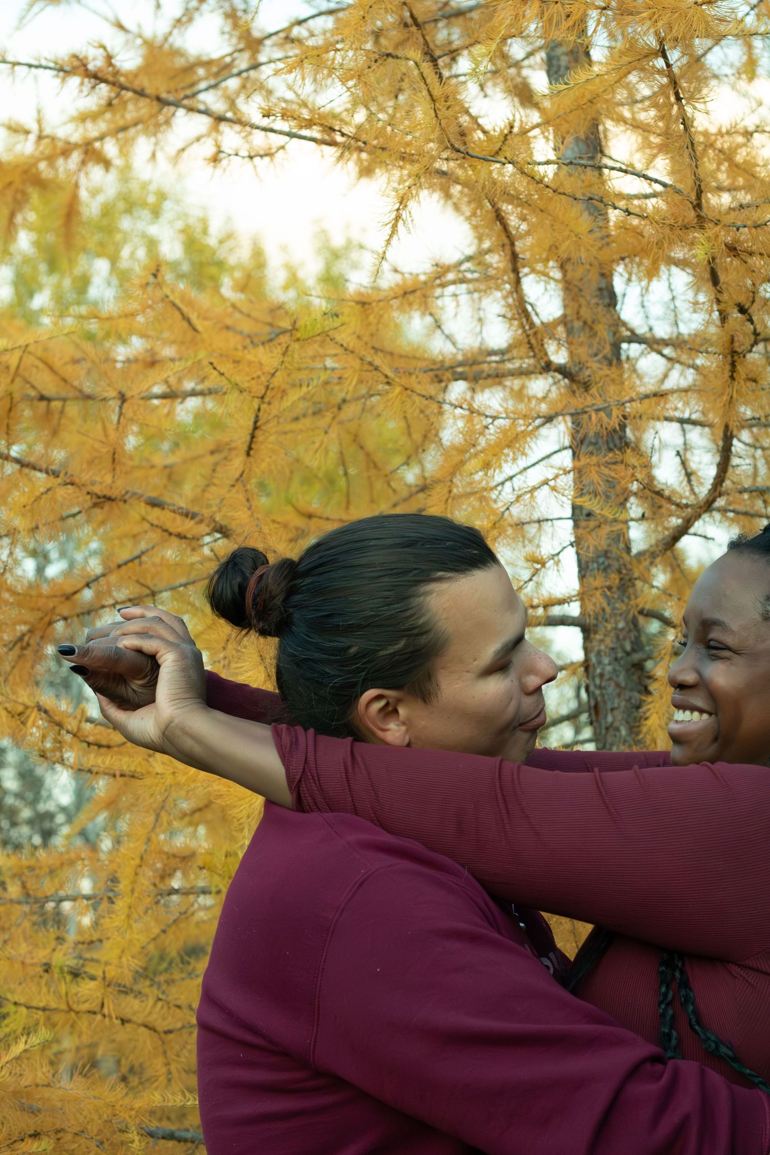 Two people in matching burgundy outfits laughing and embracing near golden larch trees in fall.