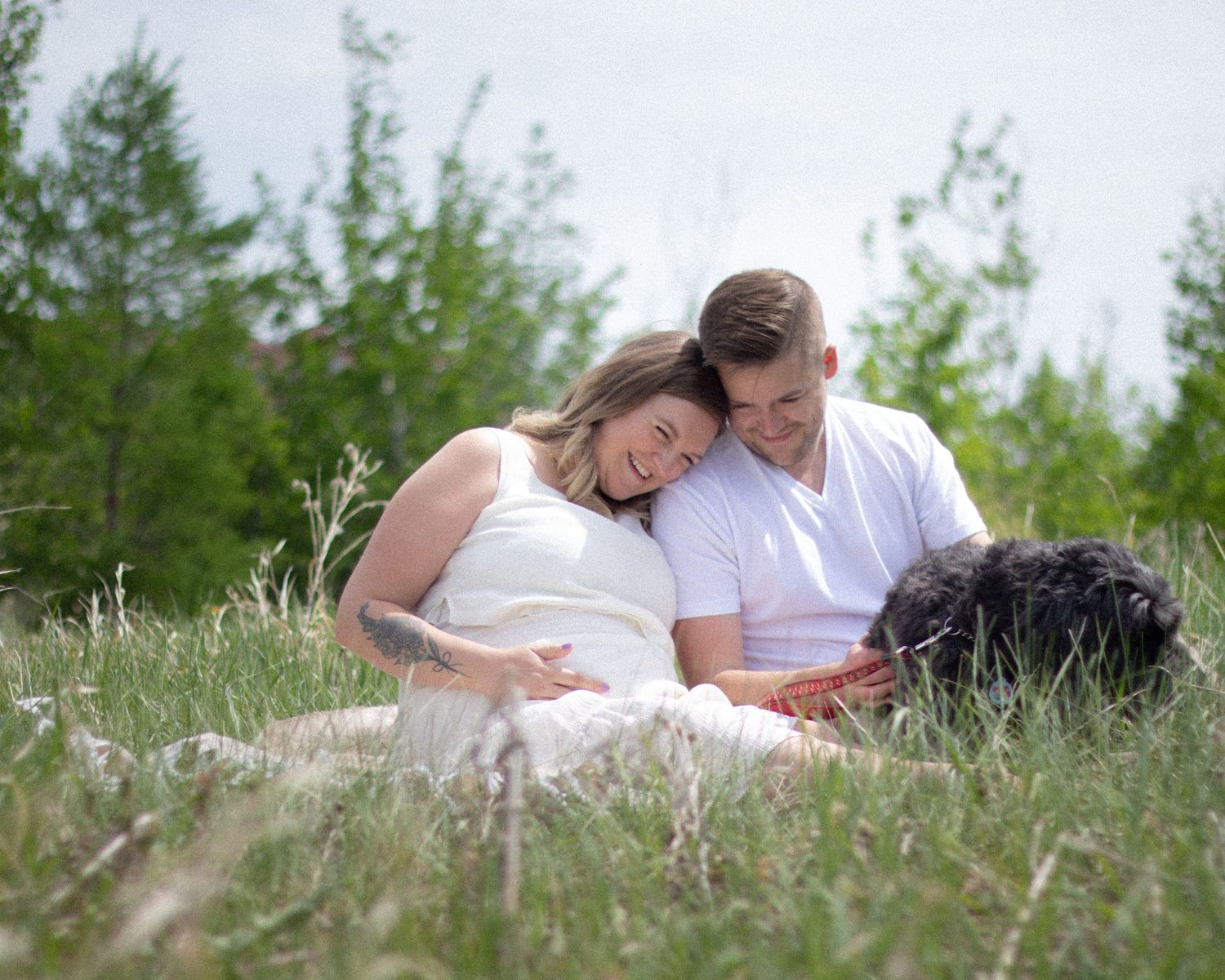 Couple relaxing in green grass field with black curly dog during outdoor maternity photo session.