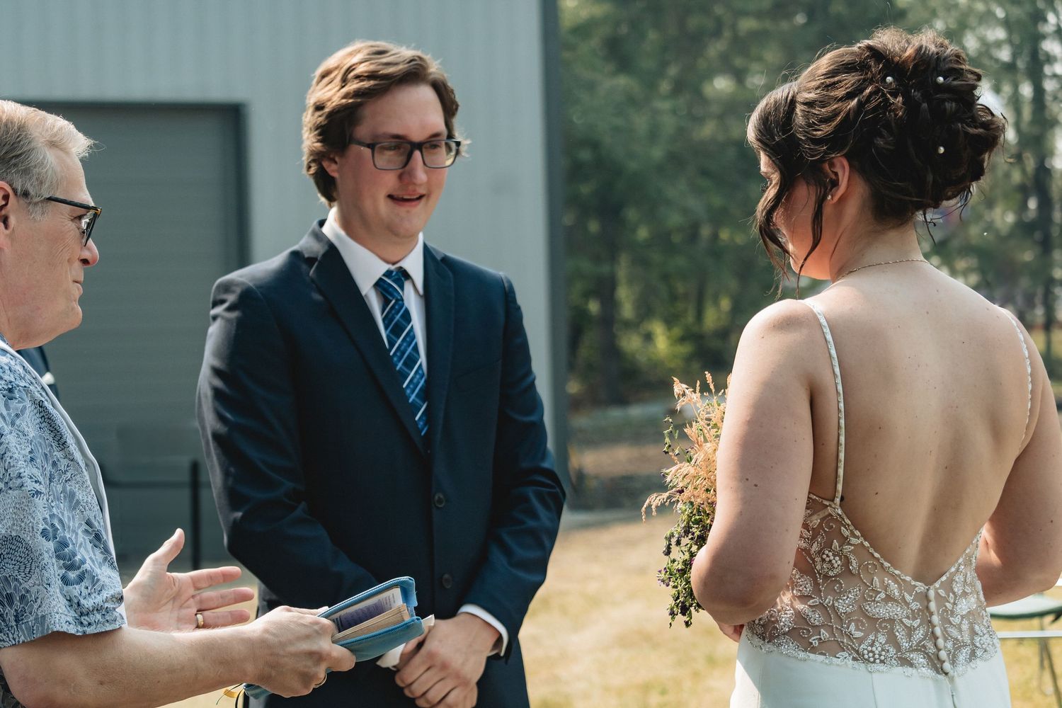 Groom smiling at bride during outdoor wedding ceremony while officiant reads from a book.