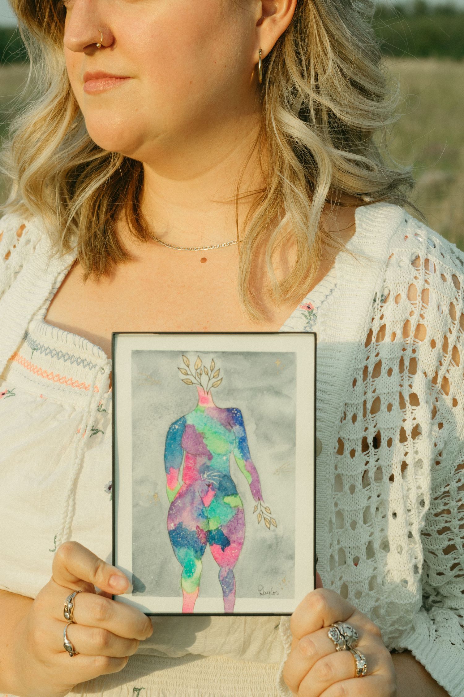 Close-up of woman holding framed colorful female figure artwork in a sunny field.