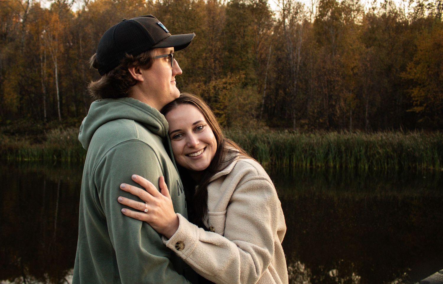 Young woman smiling while embracing her partner outdoors near a serene autumn lake.