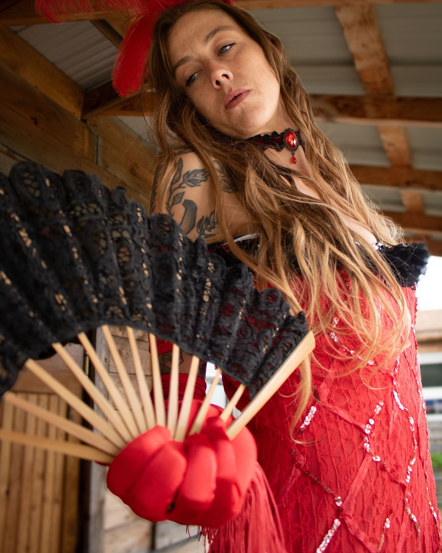 Woman in a red sequined costume holding a black lace fan in a rustic wooden setting.