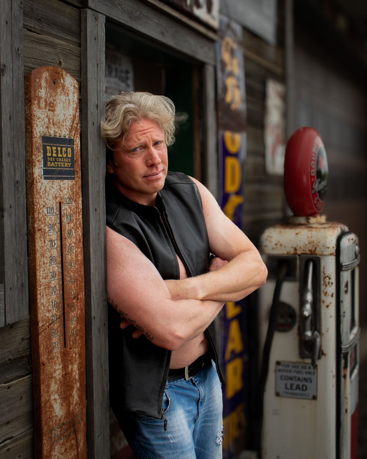 Muscular man in a leather vest leaning against a rustic wooden building by a vintage gas pump.