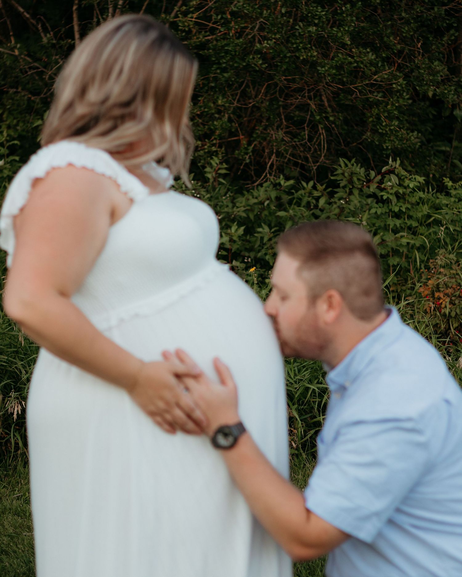 Man kneeling and kissing pregnant woman's belly outdoors in white maternity dress.