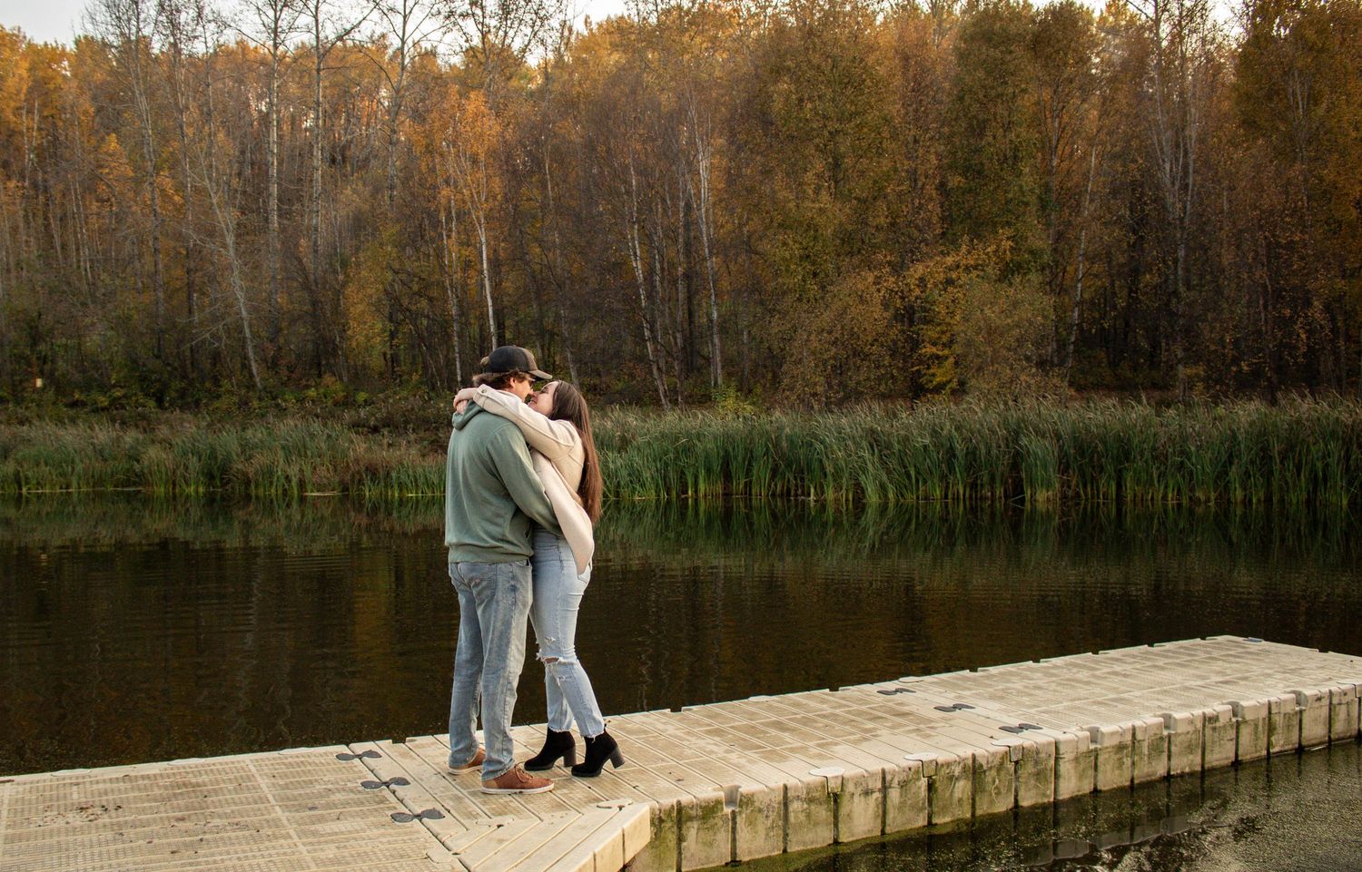 Romantic couple kissing on a wooden dock by a peaceful lake with golden autumn trees behind.