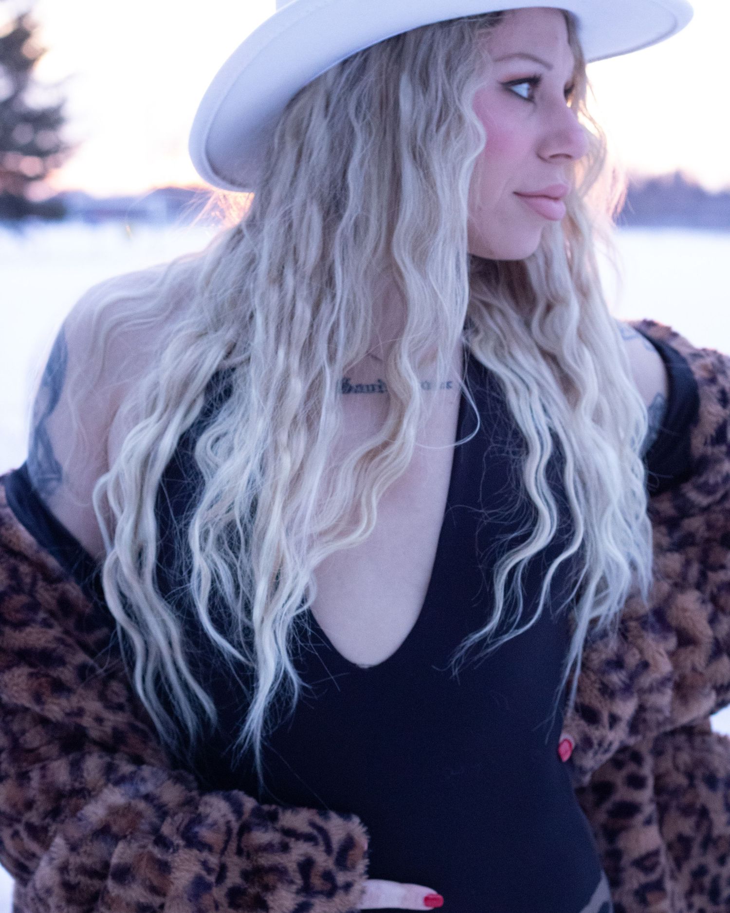 Blonde woman in white hat and leopard coat gazing sideways in snowy winter landscape at sunset.