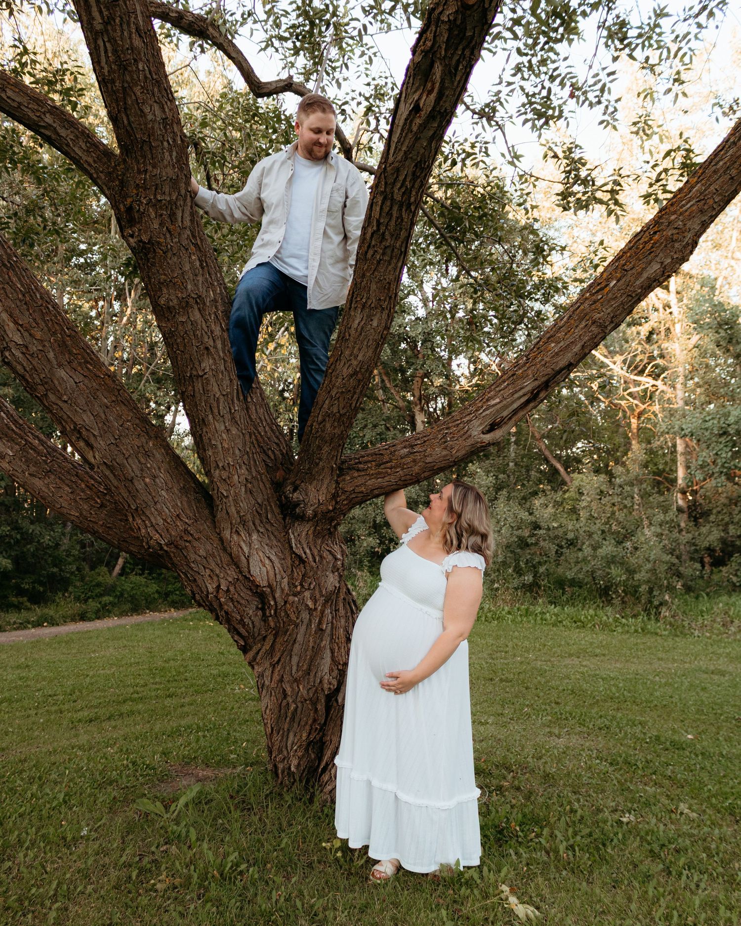 Man climbing a large tree while pregnant partner in white dress looks up smiling below.