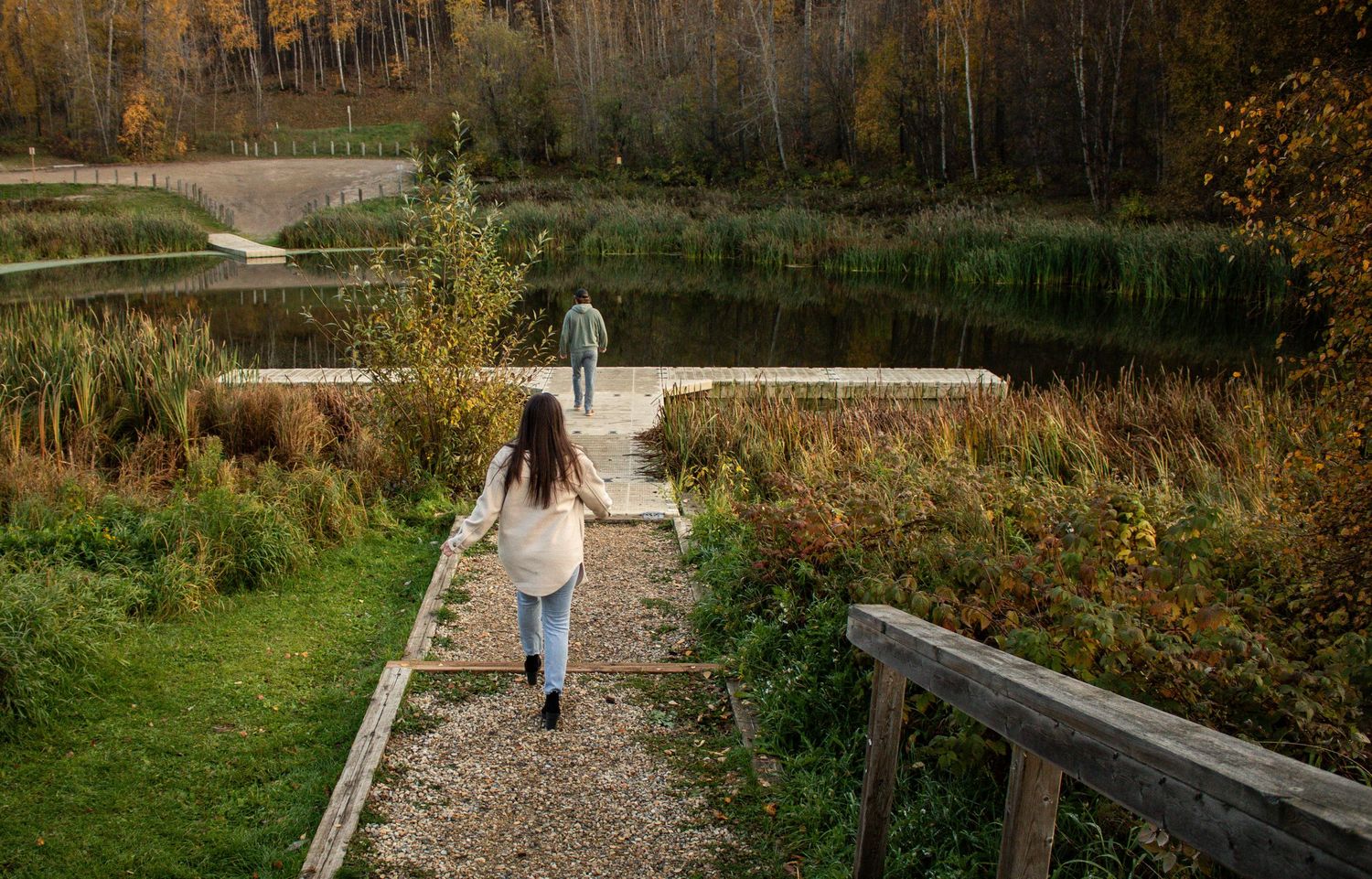 Young woman walking along a wooden path toward a serene autumn lake with colorful fall trees.