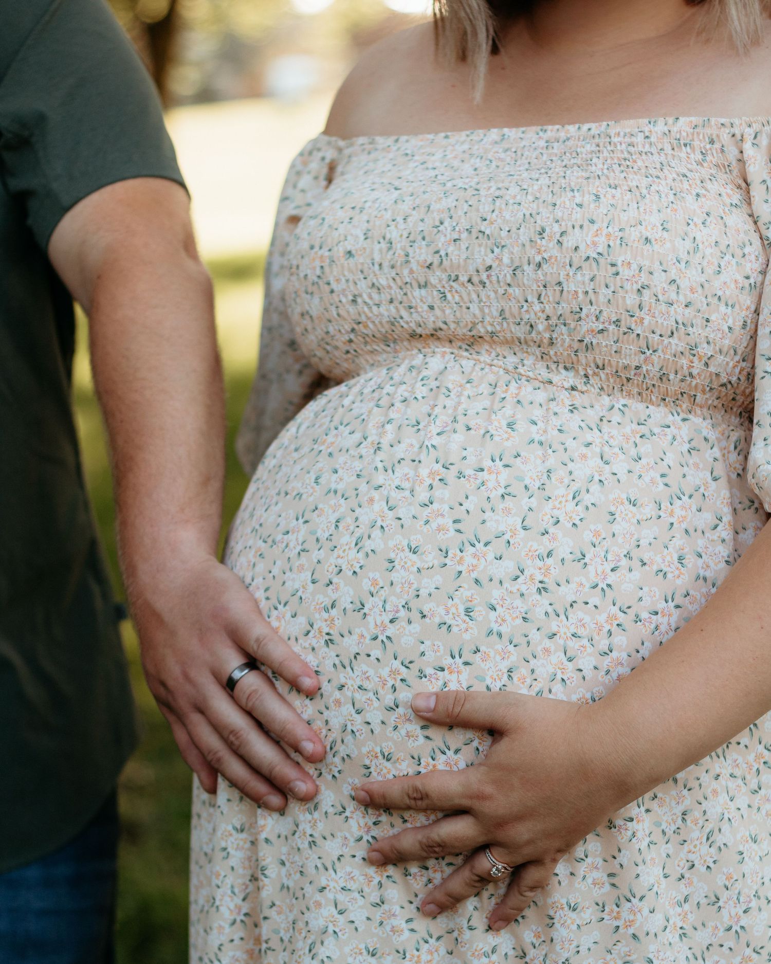Close-up of pregnant woman in floral dress with partner's hand resting on her baby bump outdoors.