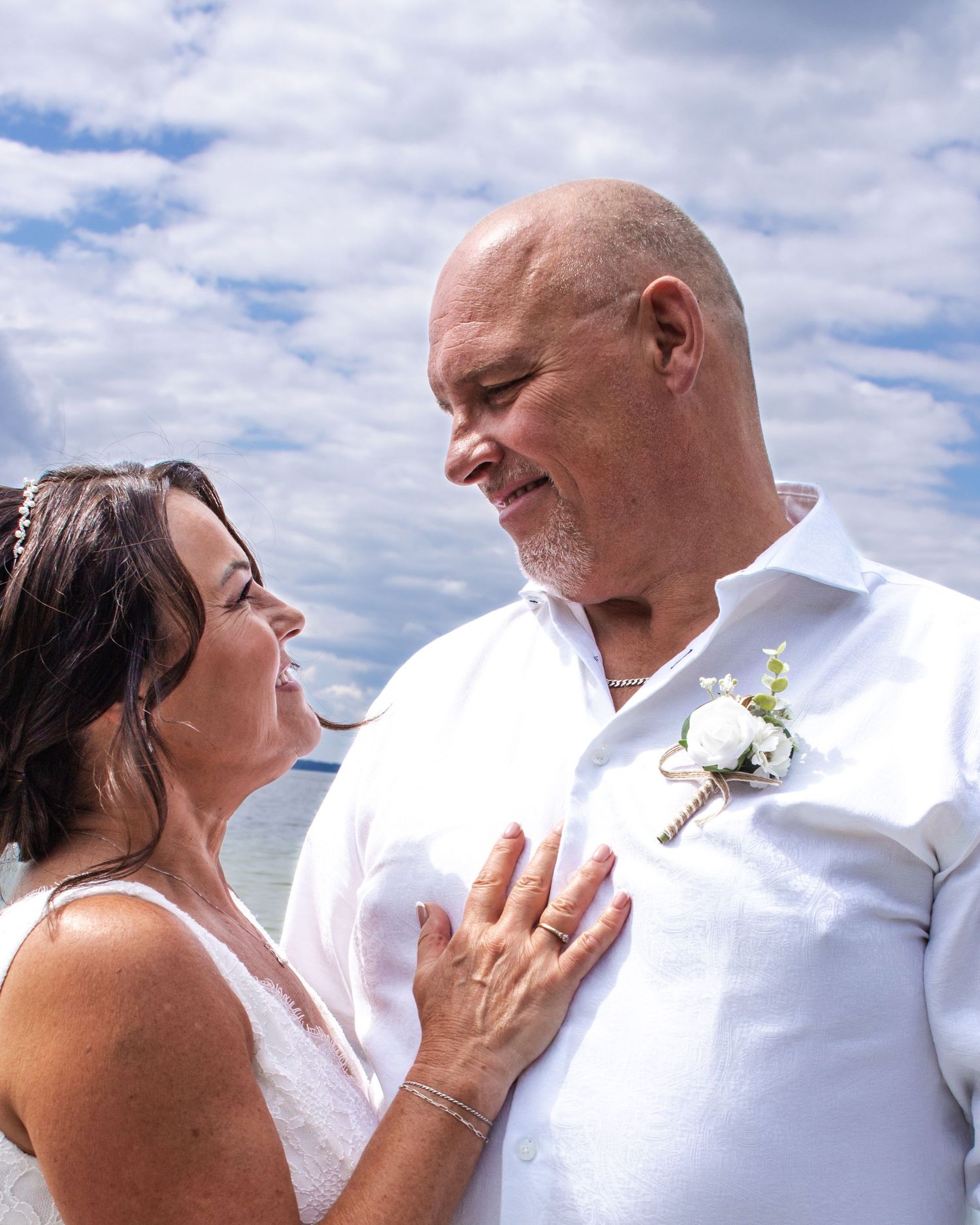Bride and bald groom share a tender moment outdoors, bride in white dress, groom wearing white boutonniere.