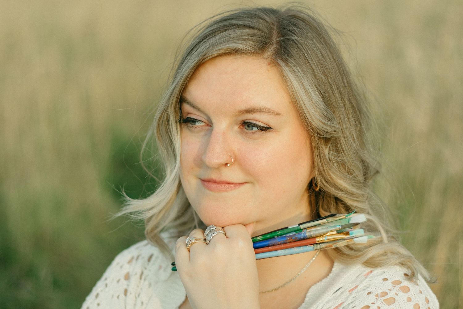 Blonde woman with nose ring holding paintbrushes under her chin in a golden field.