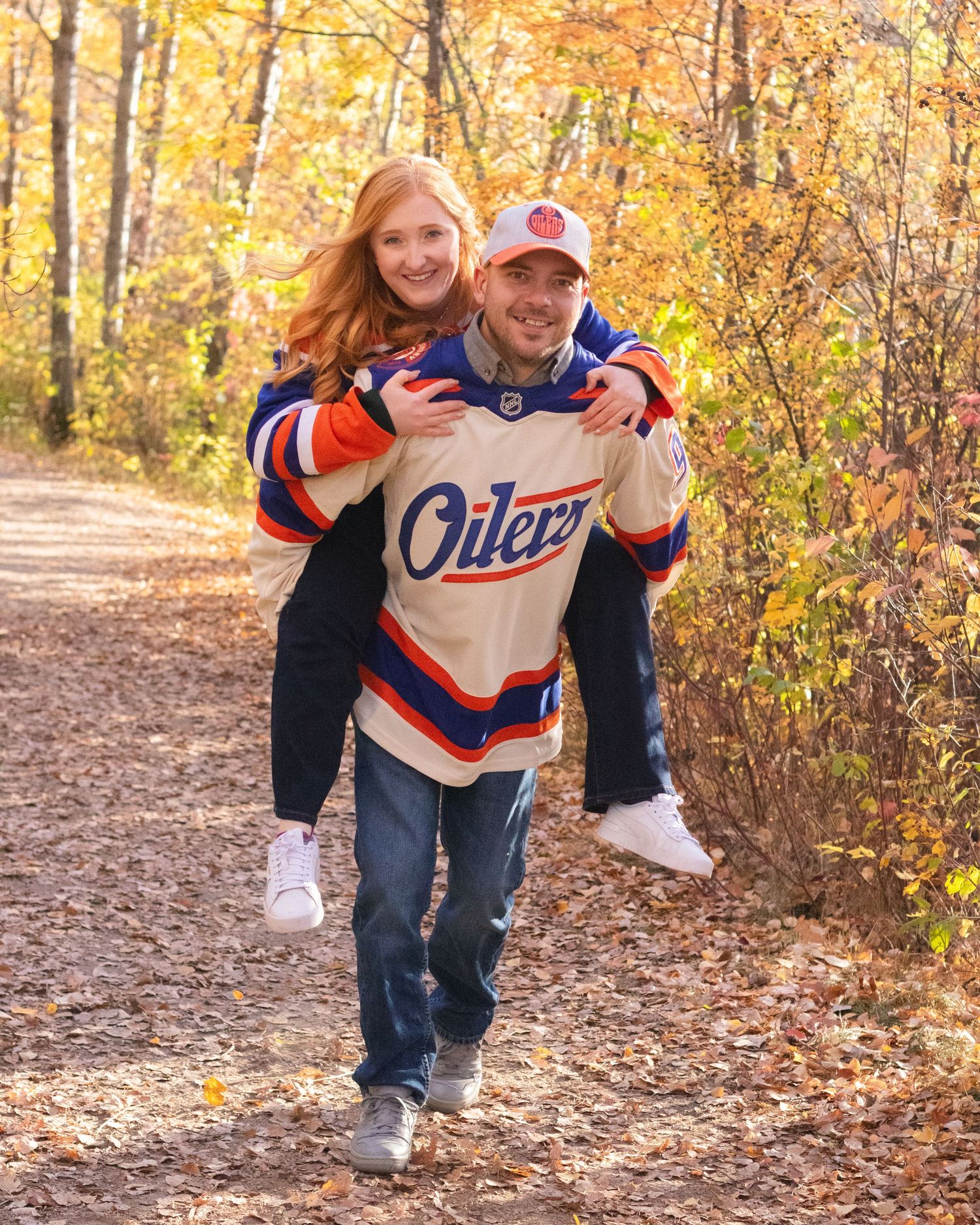 Man giving woman a piggyback ride on a trail surrounded by golden autumn foliage, wearing Oilers jerseys.